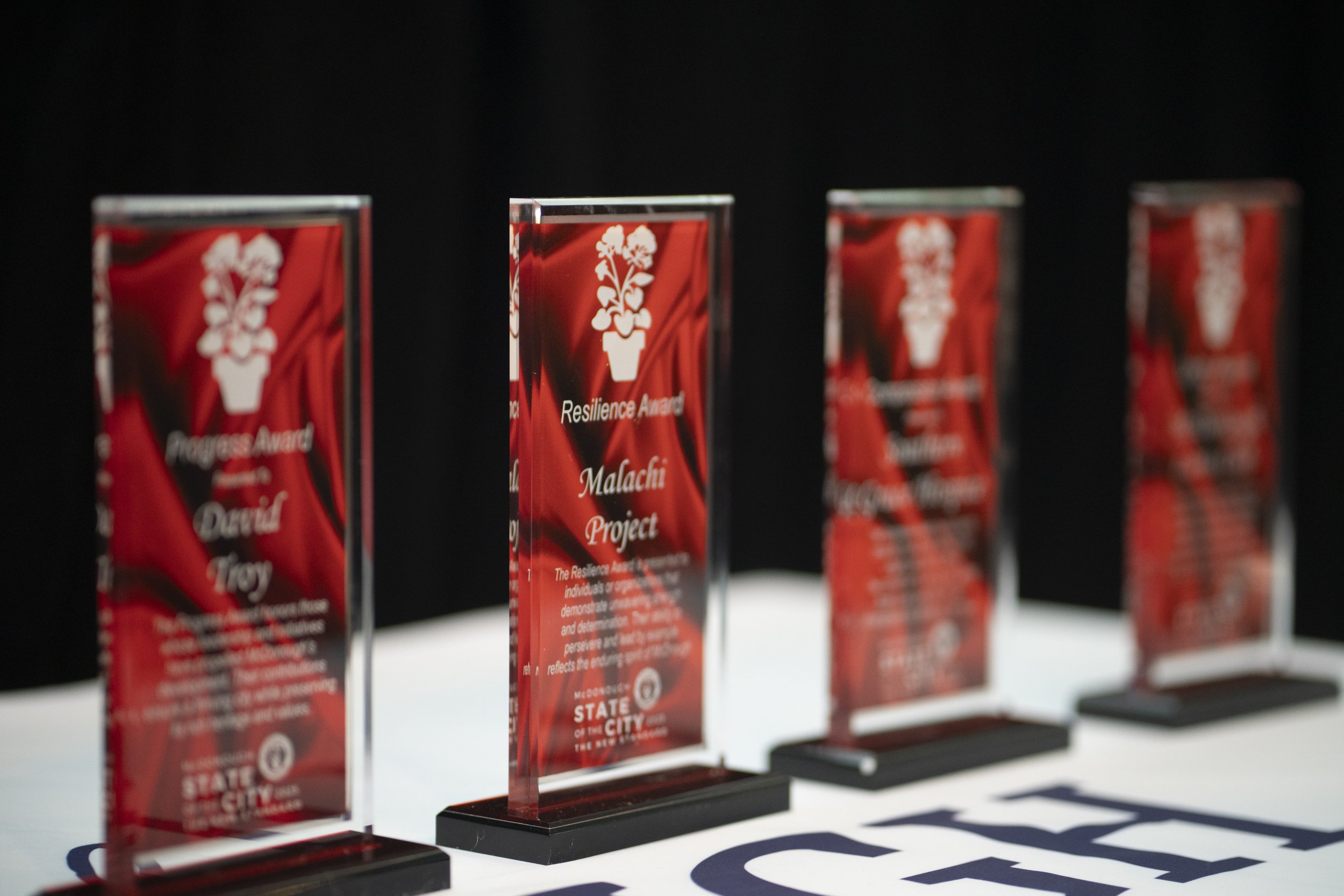 Four glass awards with red backgrounds and white text, placed on a table, with one labeled 'Resilience Award' and 'Malachi Project'.