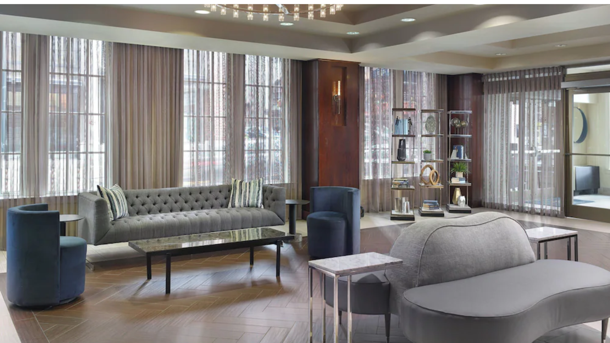 A modern hotel lobby with large windows, sheer curtains, a tufted gray sofa with striped pillows, two blue armchairs, a marble coffee table, a gray lounge chair with a marble side table, and a wooden bookshelf with decorative items. There is a sliding glass door leading to an outdoor area.