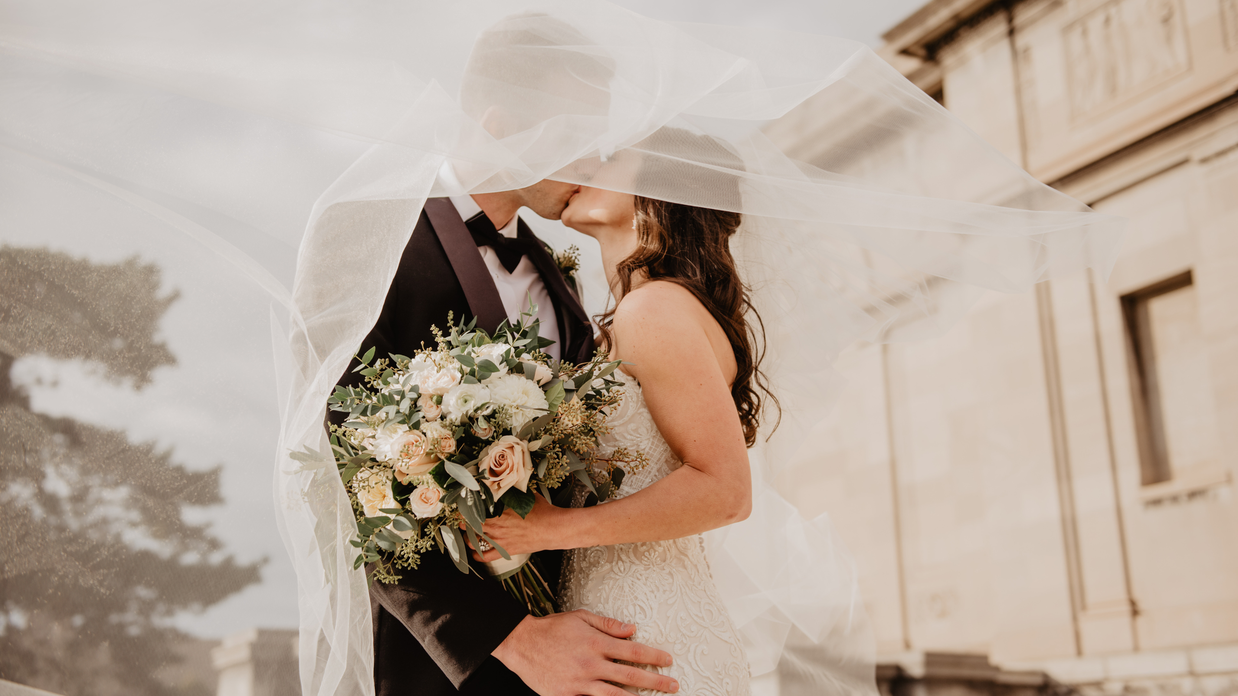 A bride and groom share a kiss under a veil. The bride holds a bouquet of white and pink flowers, and they are dressed in wedding attire outside of a building.