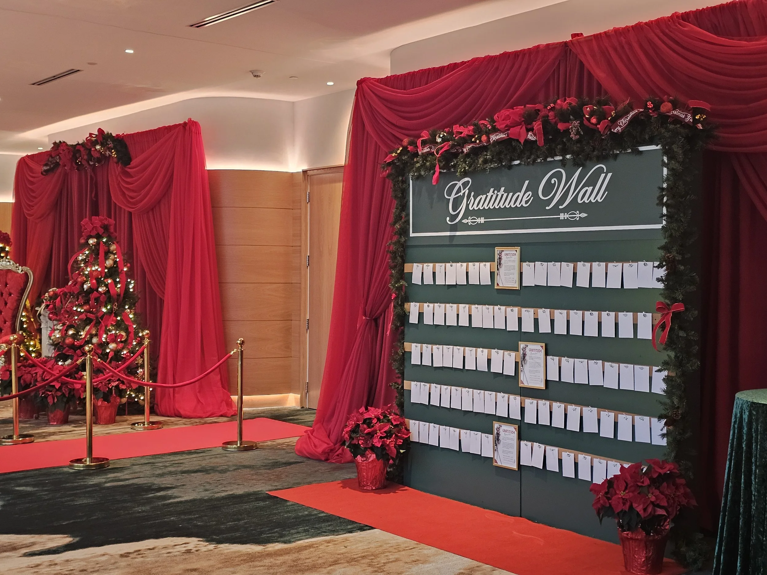 Holiday-themed gratitude wall with notes, decorated with red poinsettias, Christmas tree, red curtains, and gold stanchions.