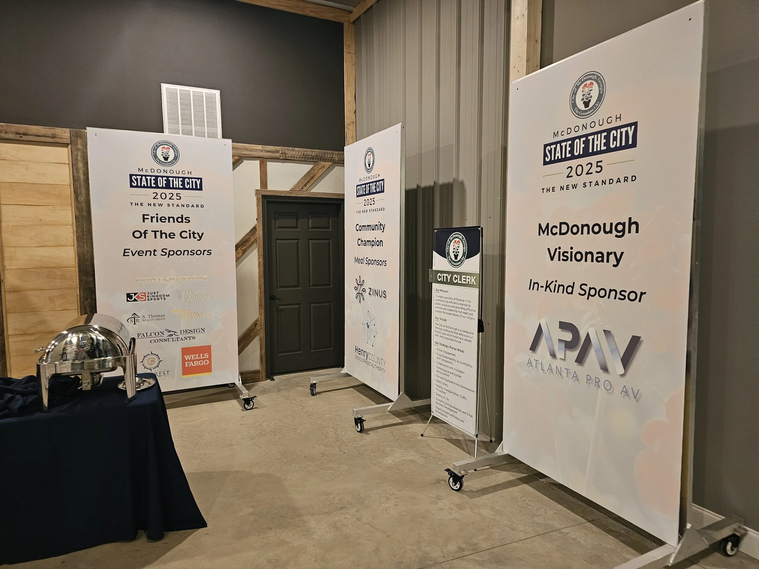 Indoor event space with three large vertical banners displaying sponsor information, a table with a cloche cover, and a closed black door. The banners feature logos and text for the McDonough 'State of the City 2025' event, including sponsors like We