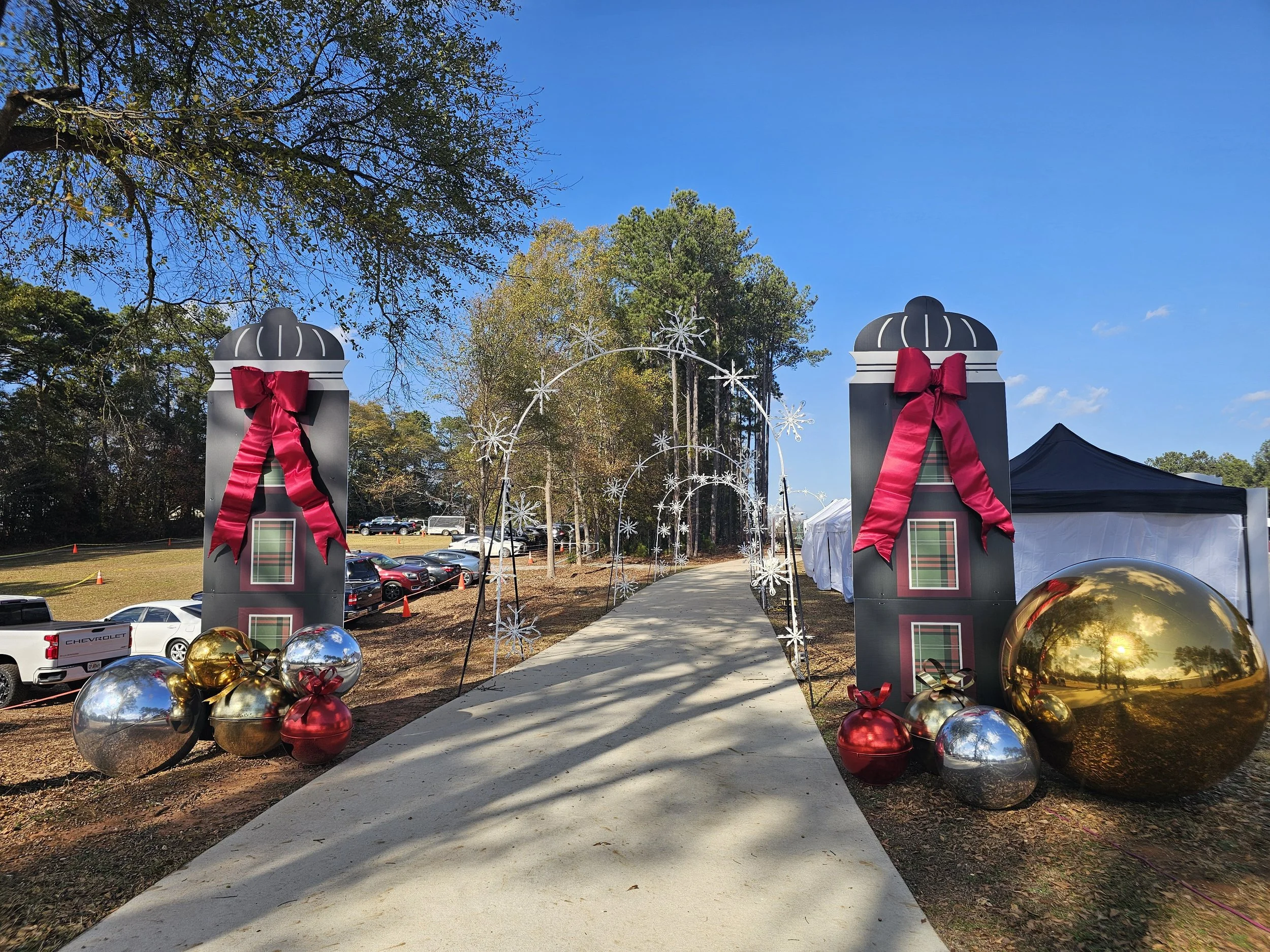 Decorative holiday display with oversized Christmas presents, bows, and ornaments on either side of a pathway under festive arches, set outdoors on a sunny day with trees and parked cars in the background.