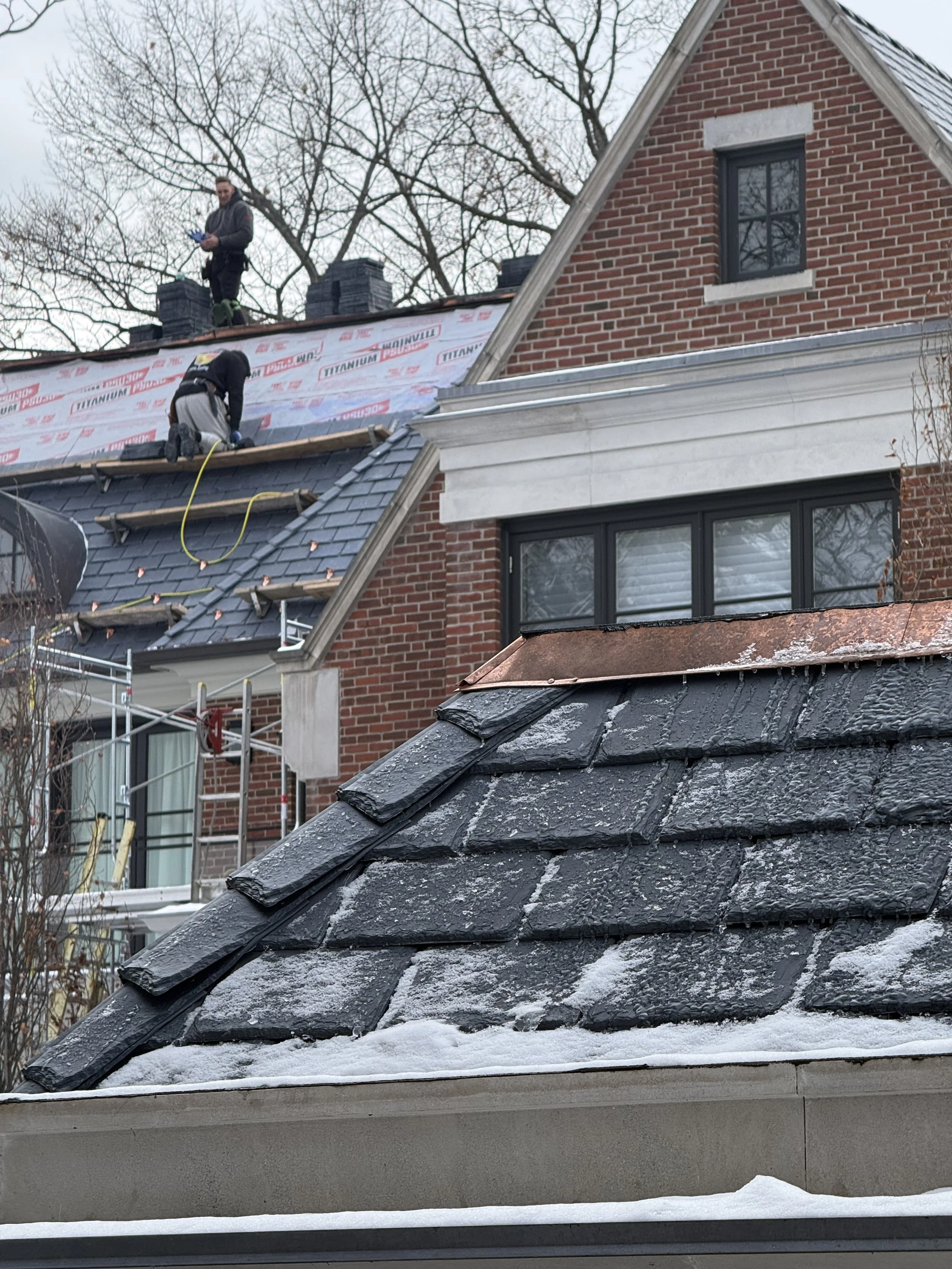 Two workers on a rooftop under construction, with one kneeling on the shingles and another standing on the roof, partial snowy house scene with a brick house, leafless trees, and construction materials.