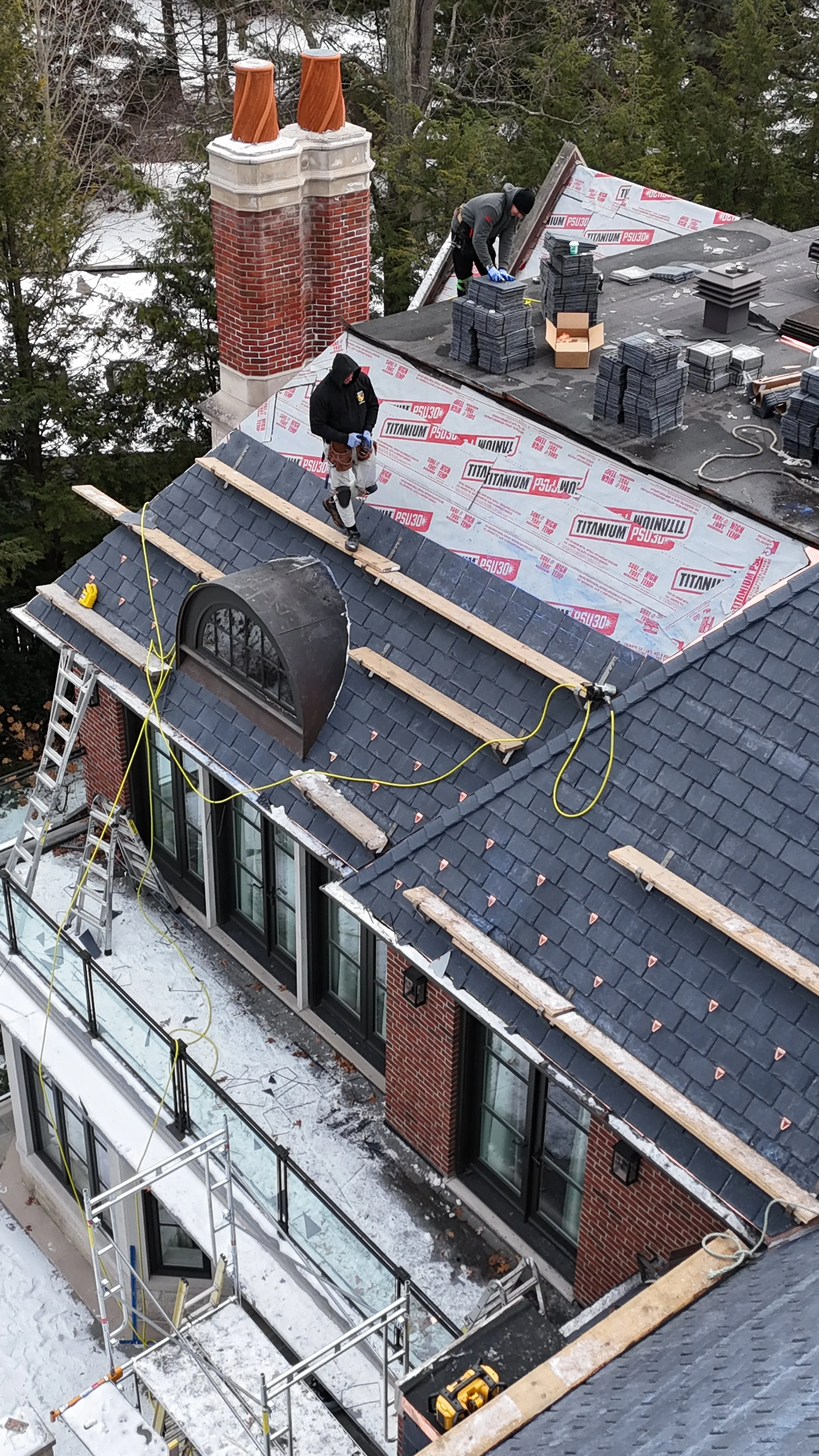 Workers installing or repairing roofing on a building, with stacks of roofing materials and tools on the roof, and snow on the ground.
