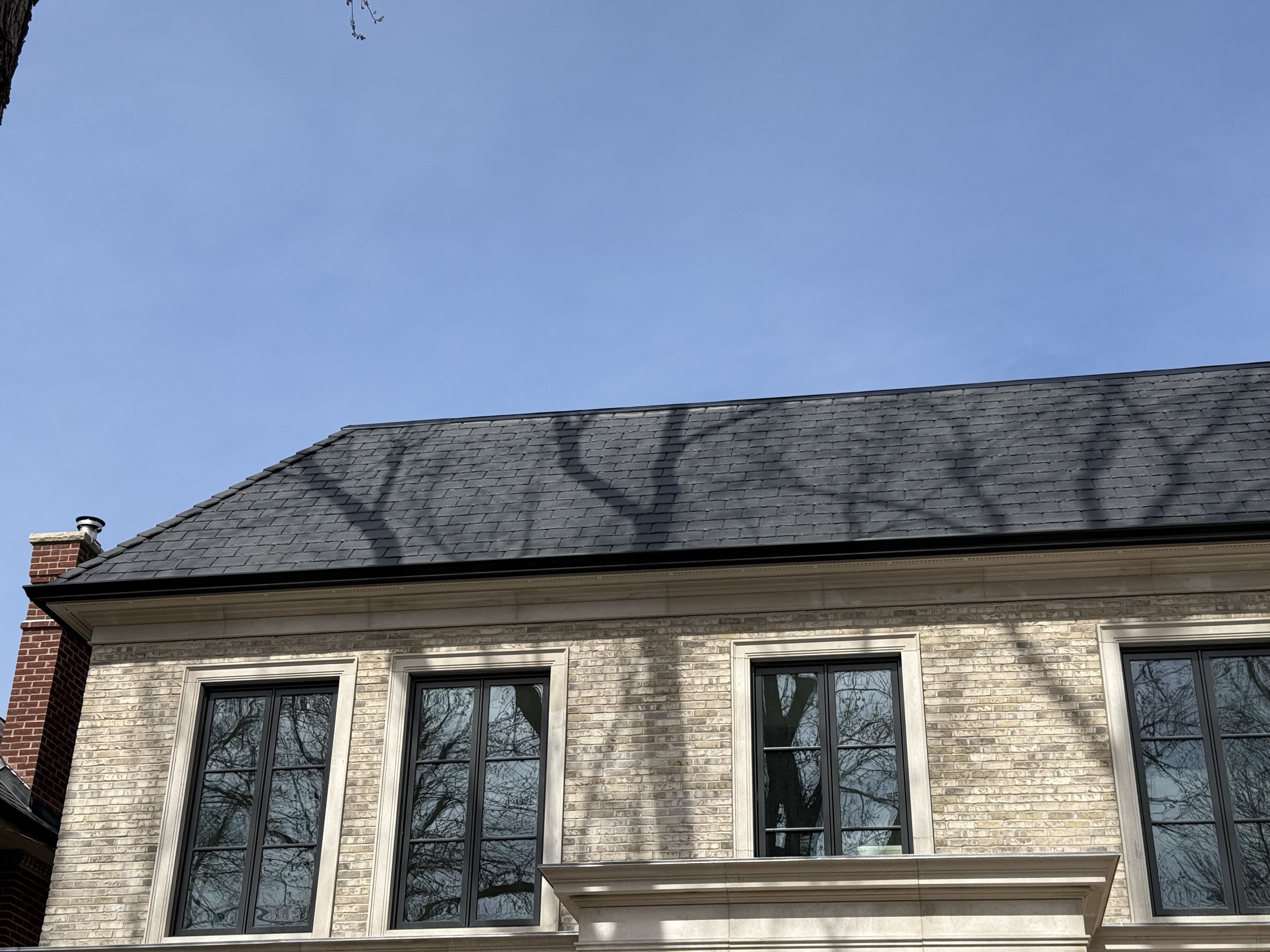 Shadow of tree branches on the roof of a brick house with gray shingles and window reflections showing bare trees, under a clear blue sky.