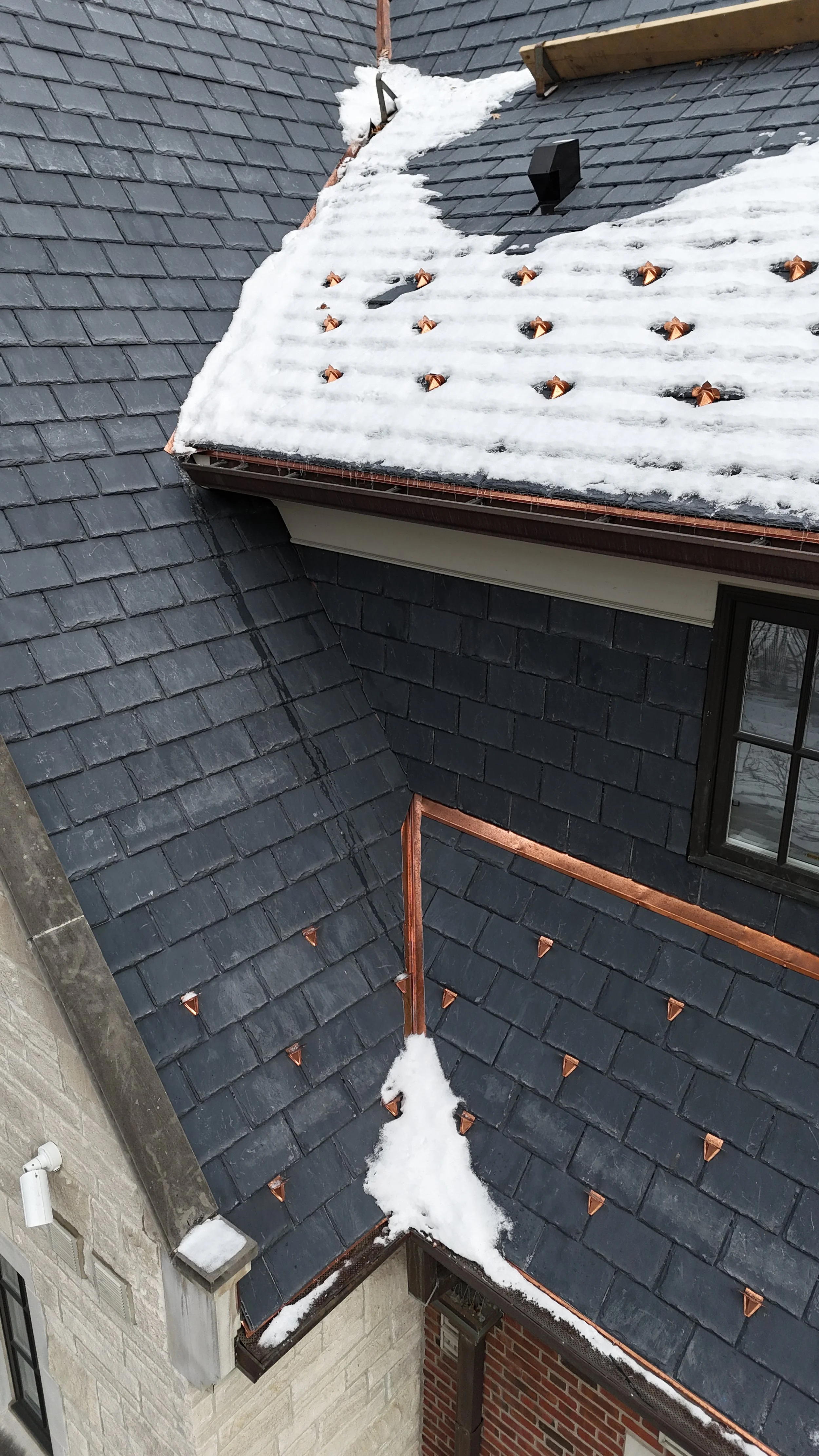 A view of a residential rooftops intersection with snow on the shingles and copper flashing, copper roof ornaments, and a brick wall with an outdoor light fixture.