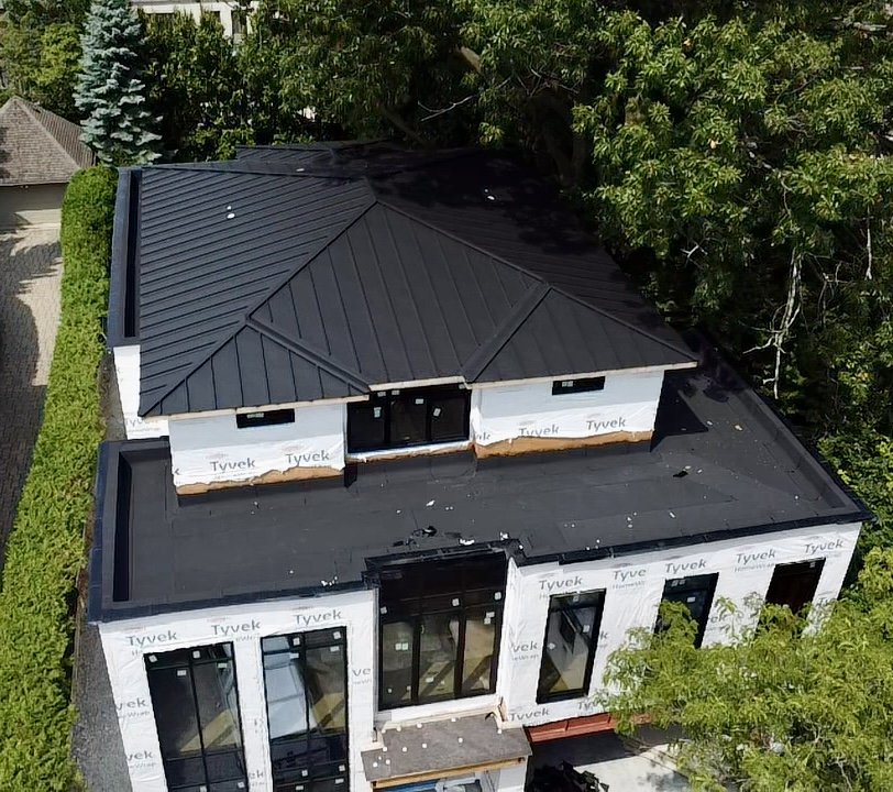 A two-story house under construction with a black metal roof, white exterior walls, and large glass doors and windows, surrounded by trees and greenery.