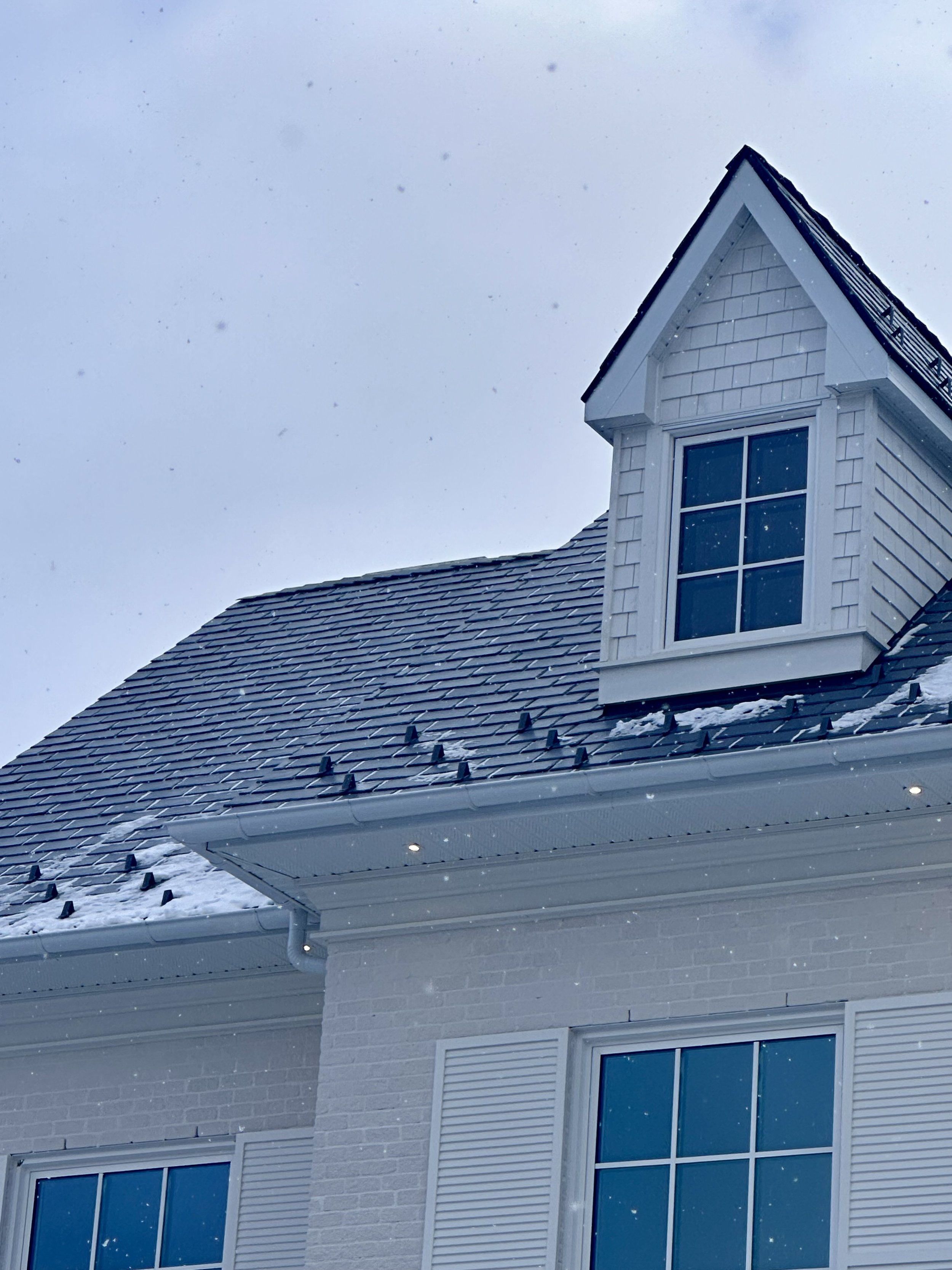 Close-up of a white house's roof and dormer window covered in snow, with a cloudy sky in the background.