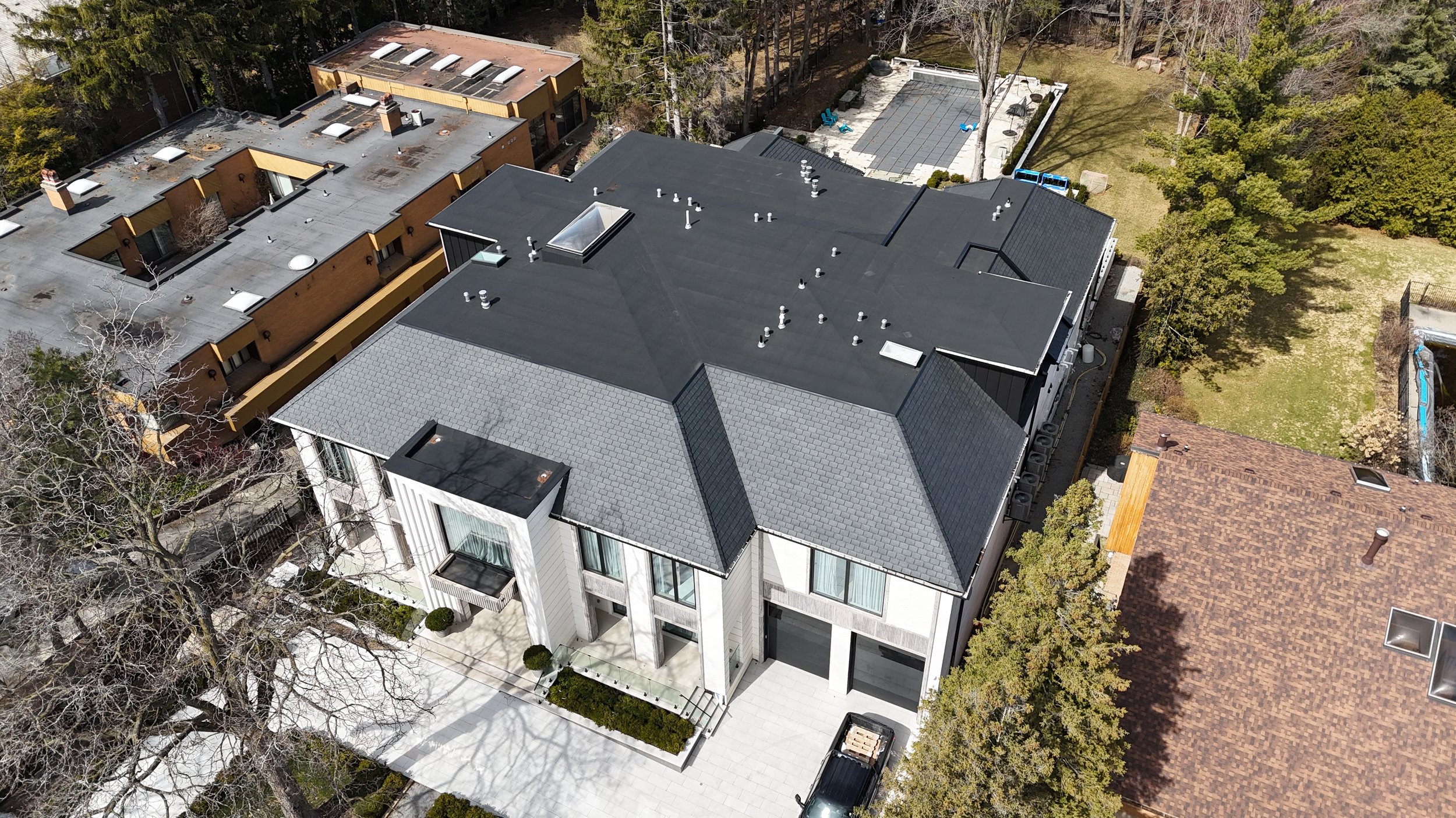 Aerial view of a modern multi-story house with a black and gray roof, located in a neighborhood with other houses, trees, and a backyard patio area.