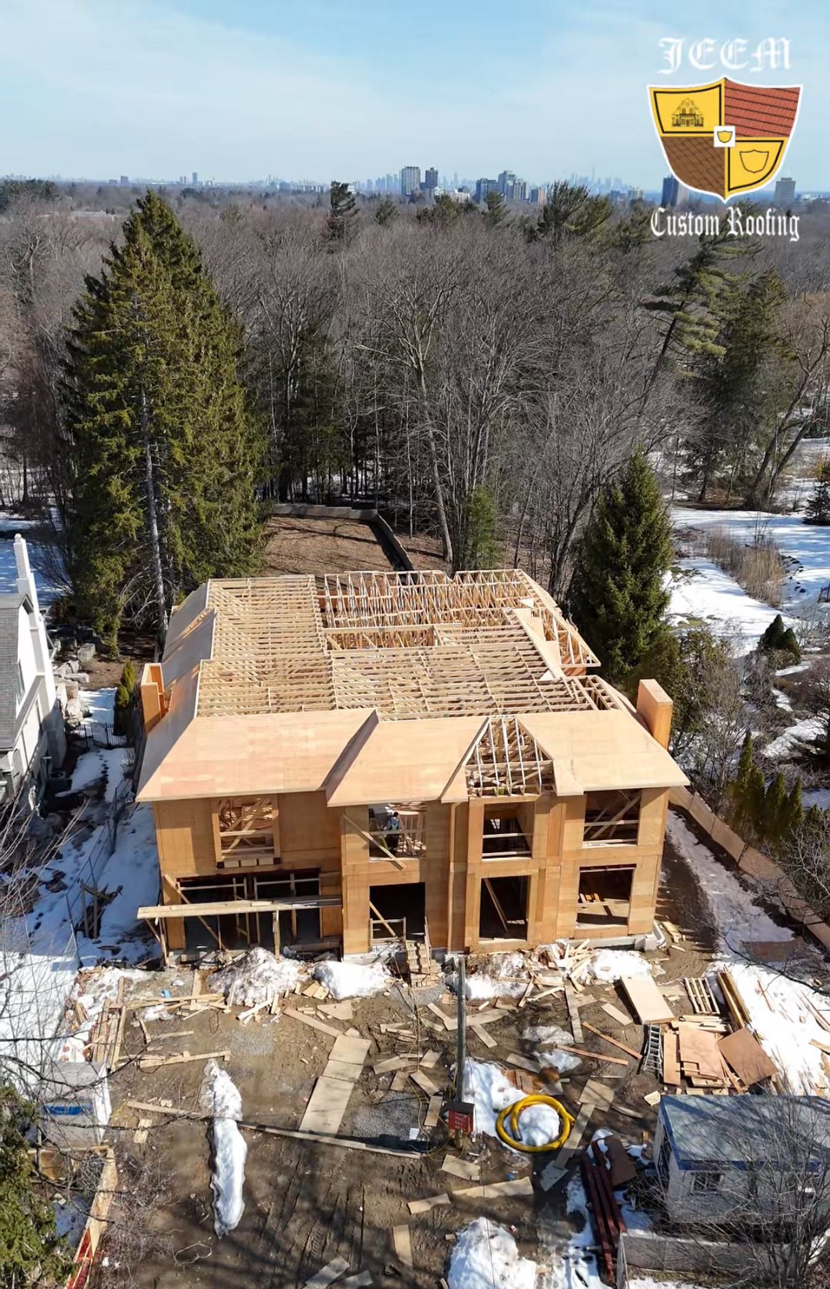 A two-story wooden house under construction in a snowy, wooded area with trees and city skyline in the background.