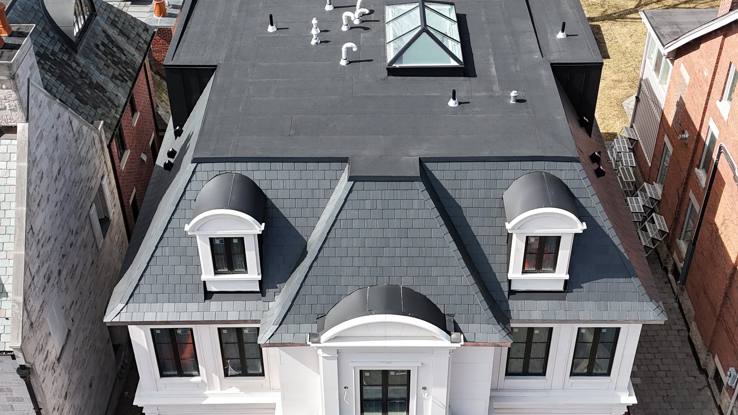 Aerial view of a building with a flat rooftop, featuring three dormer windows with arched tops and black roofs, and ventilation pipes. The building has white walls and is surrounded by neighboring brick buildings.