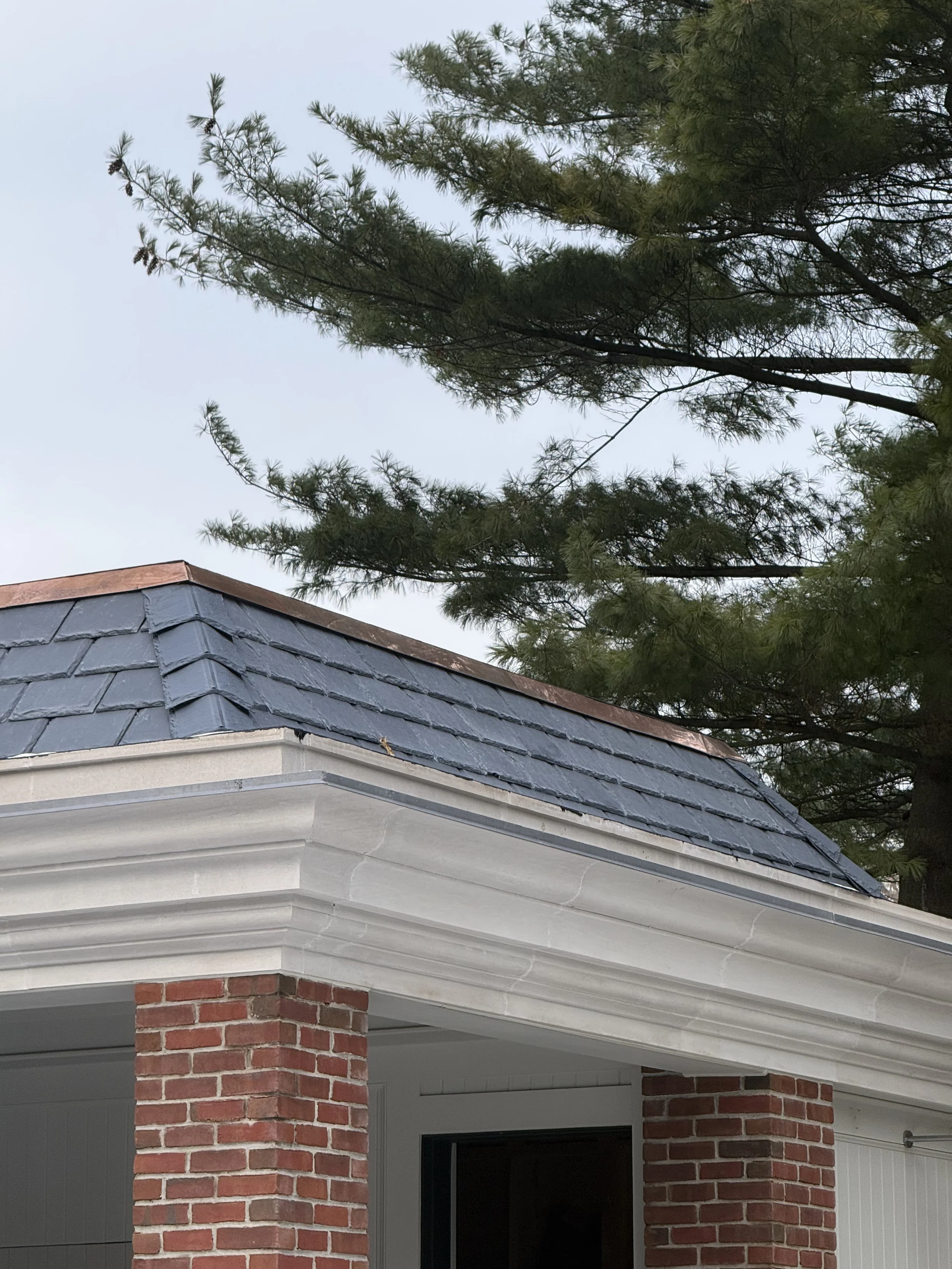 Partial view of a house with a brick pillar, white siding, and a roof with slate tiles. A large pine tree is in the background against a clear sky.