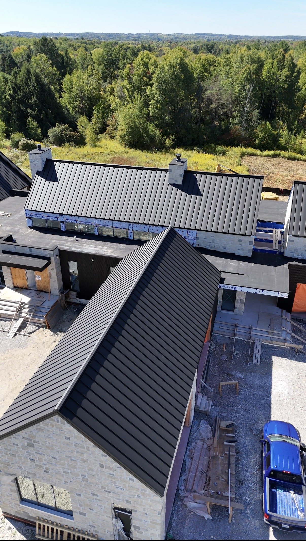 An aerial view of a construction site showing buildings with metal roofs, surrounded by trees and forest in the background.