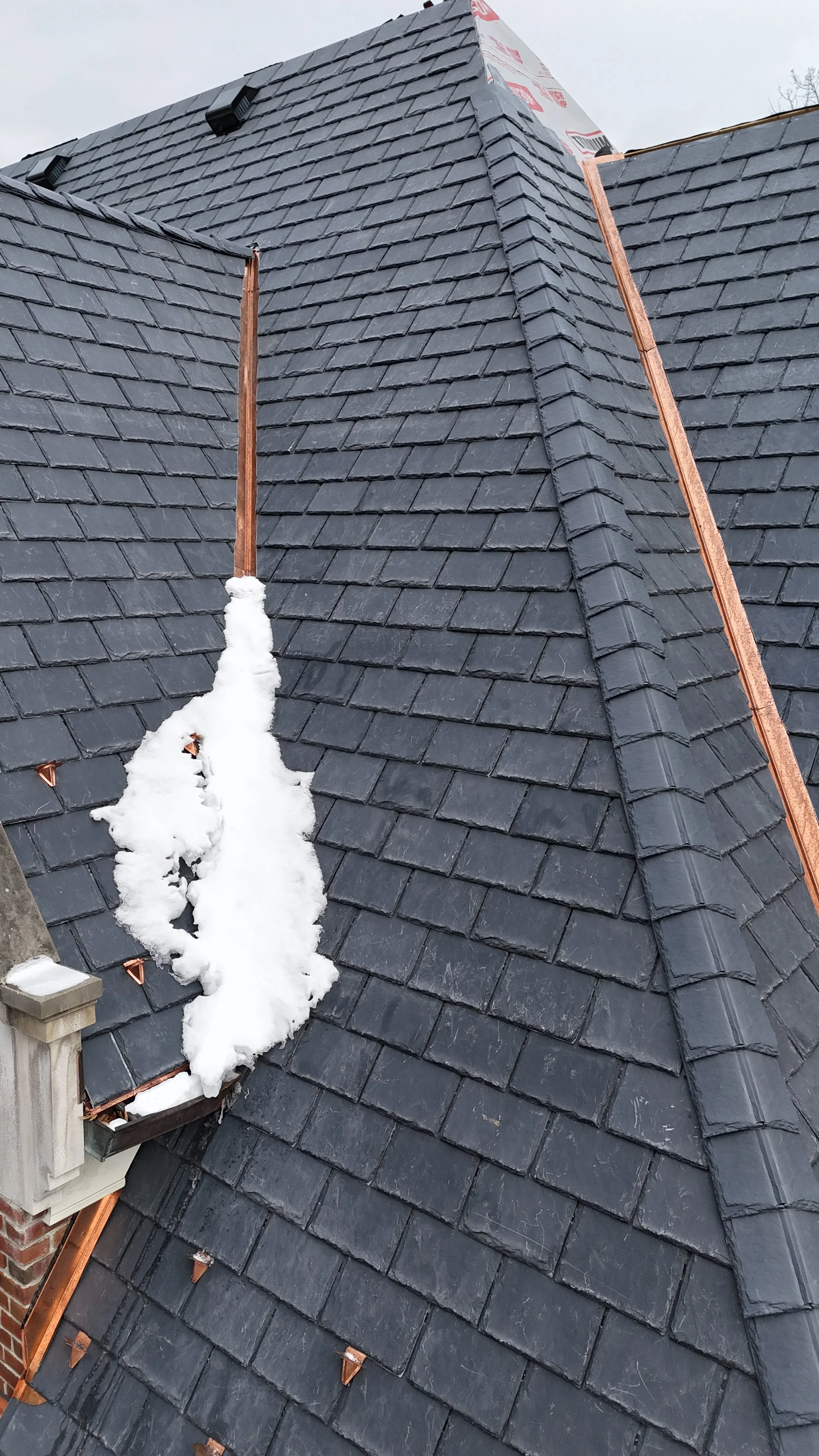 View of a black tiled roof with some snow accumulated along the edge and copper vent pipes, with a small section of brick chimney in the lower left corner.