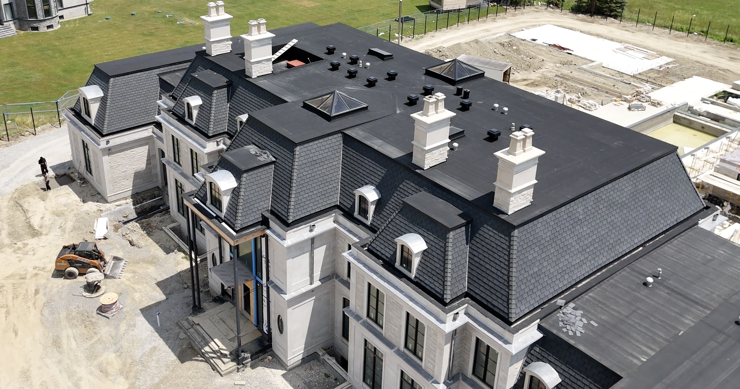 A large, multi-story building under construction with a black roof, multiple chimneys, and several dormer windows. Construction equipment and workers are visible around the building.