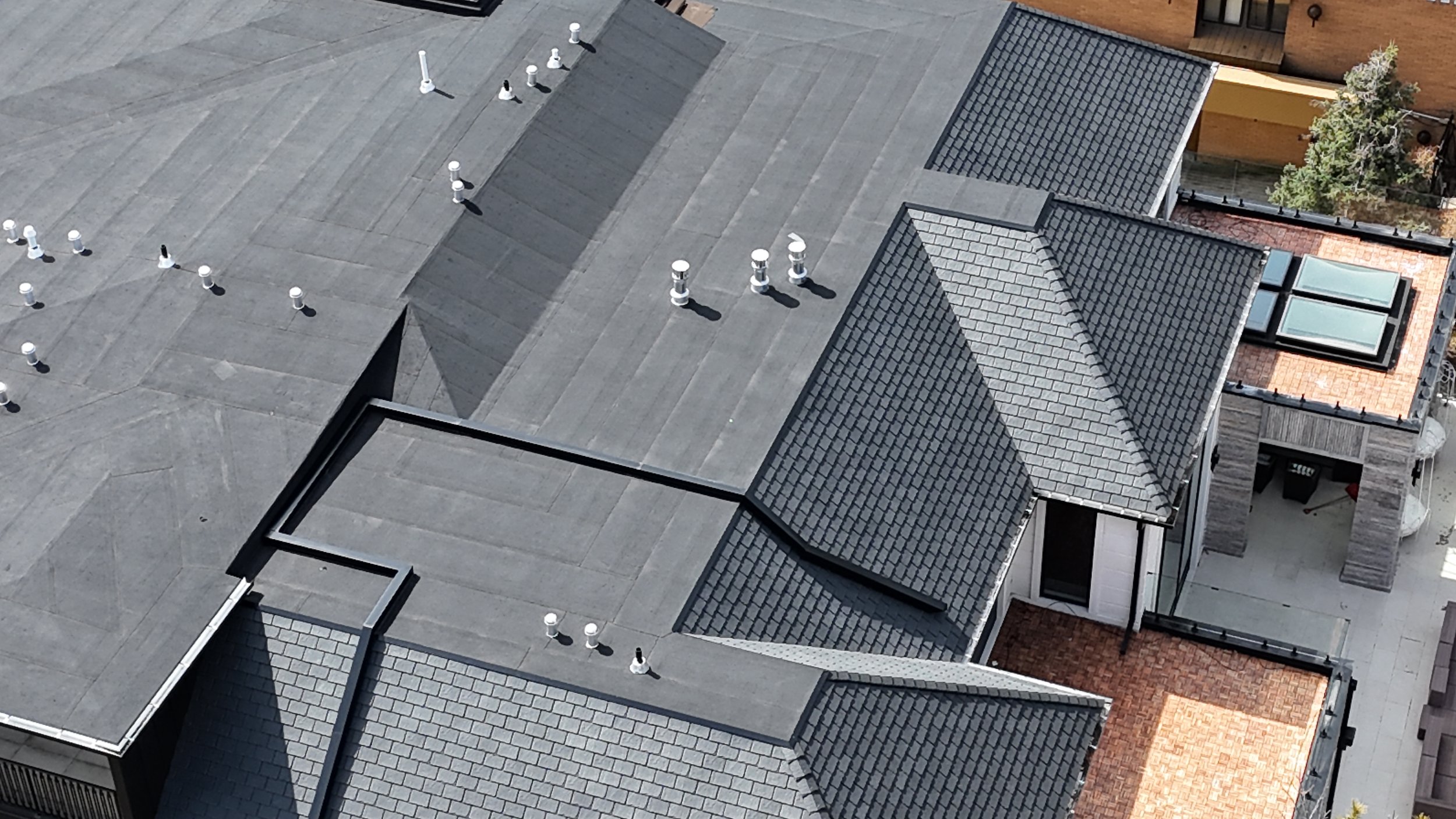 Aerial view of a rooftop with multiple gray sections, some with black vents, a glass skylight, and partial view of neighboring wooden and brick structures.
