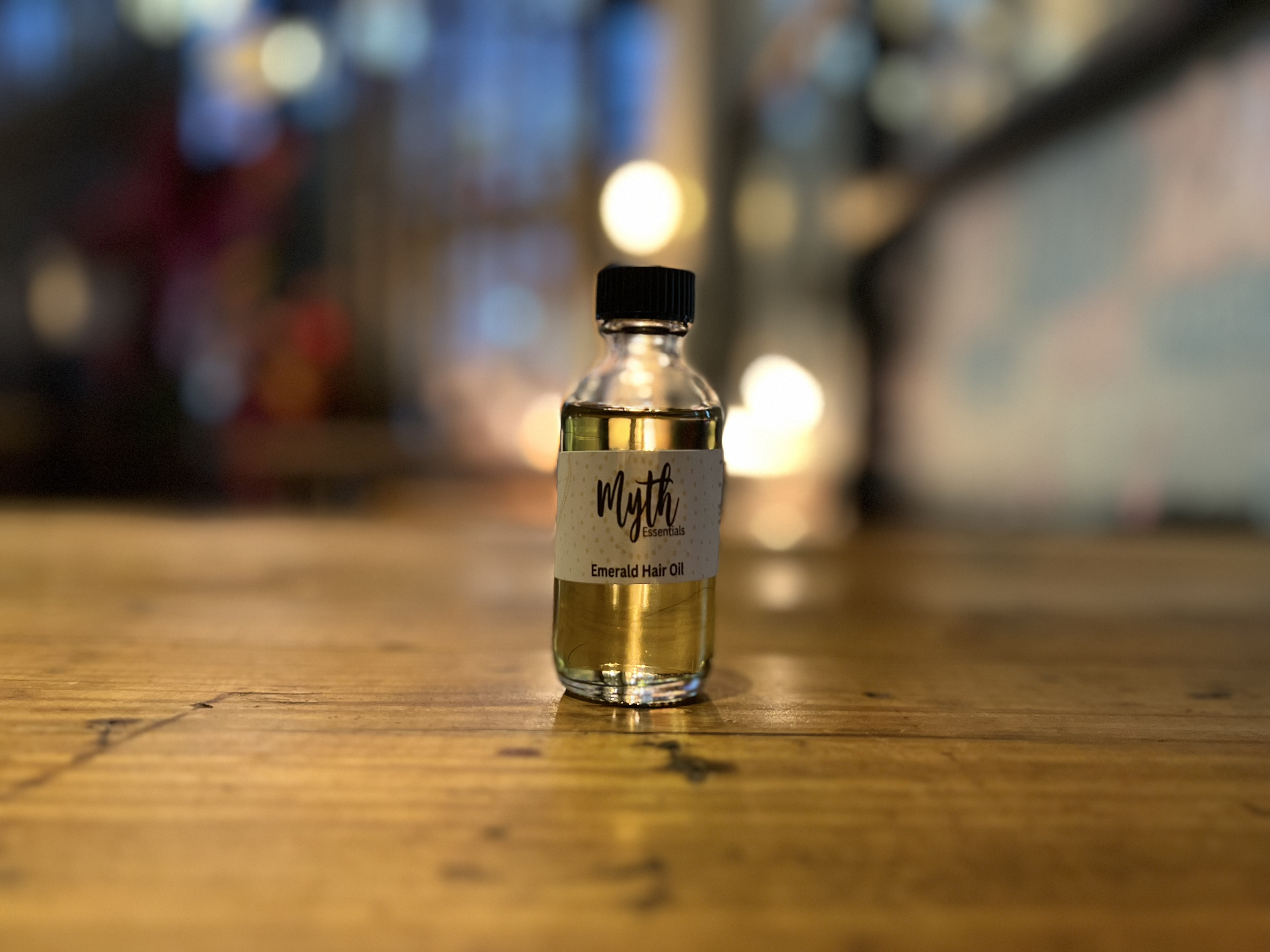 A small clear glass bottle with a black cap sitting on a wooden surface, labeled "Myth Essentials Emerald Hair Oil". The background is blurry with various lights and colors.