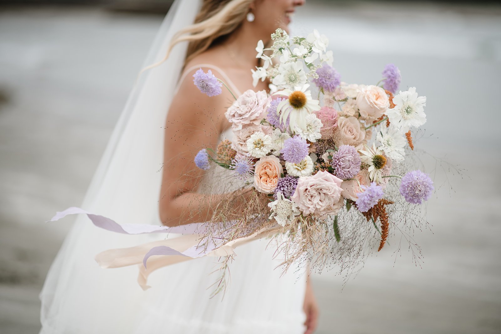 Bride holding a bouquet of pastel flowers, including lavender, peach, and white blooms, with a ribbon, outdoors.