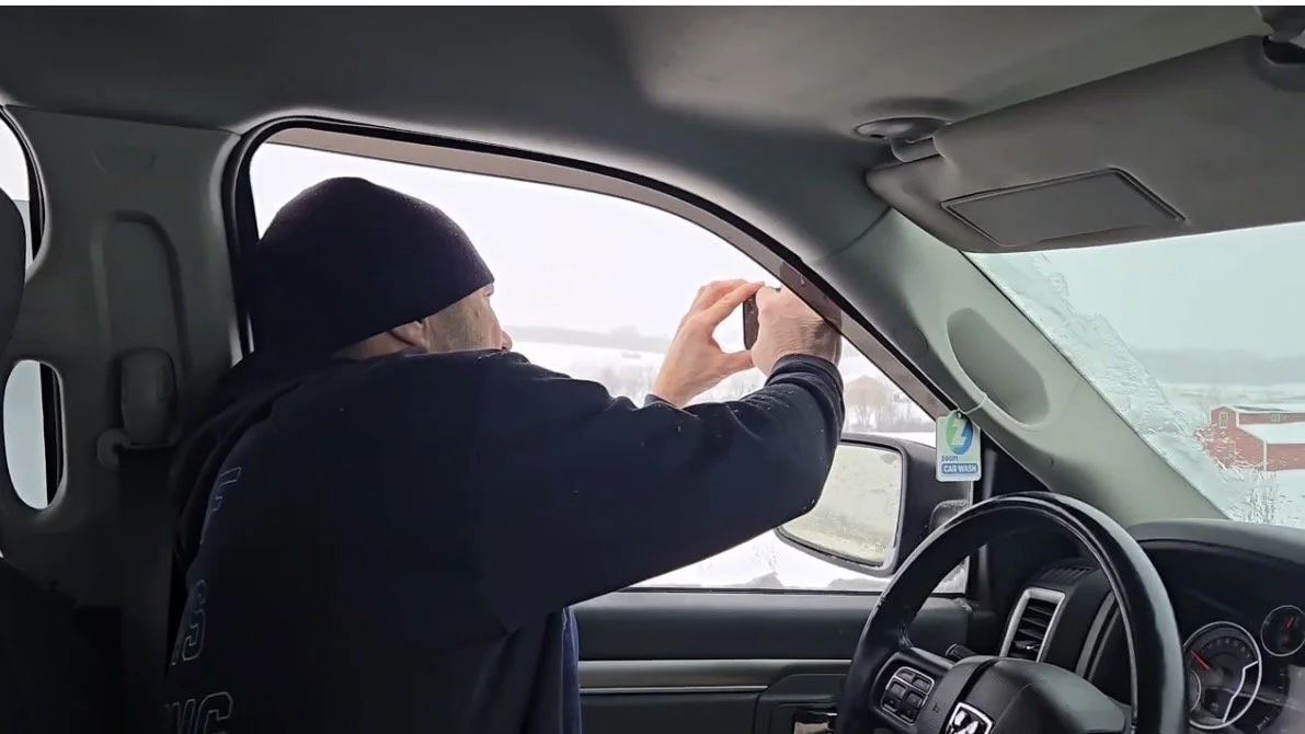 Photographer wearing a black beanie taking a photo out of the front passenger window of a vehicle during snowy weather.