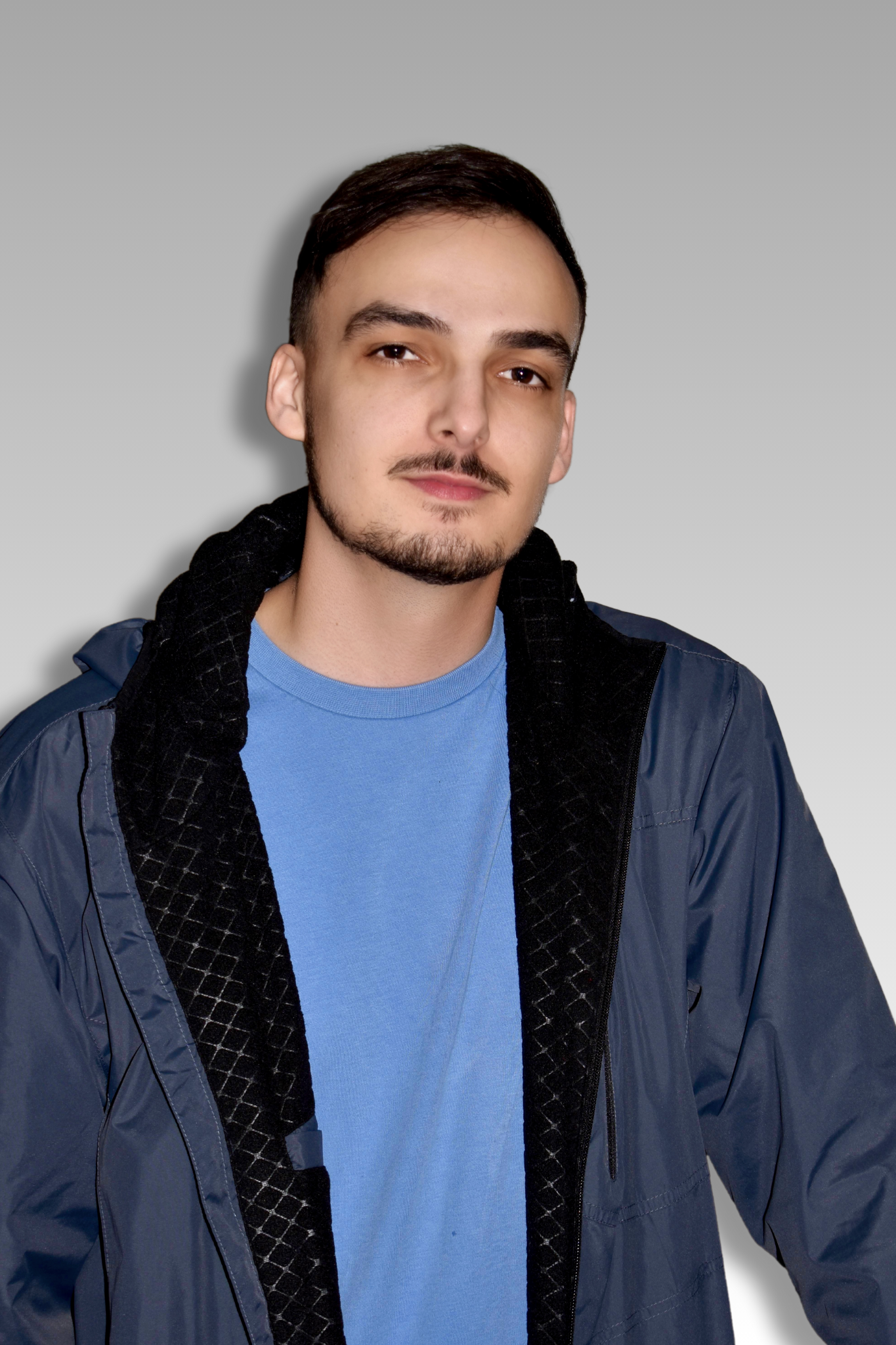 A young man with dark hair, facial hair, wearing a blue t-shirt, and a dark jacket, standing against a gradient background.