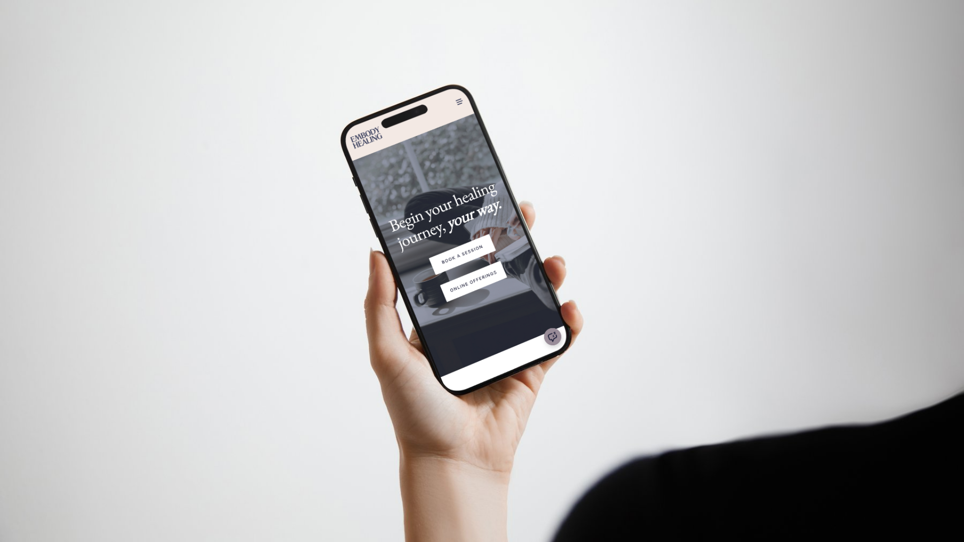 Hand holding a smartphone displaying a website with the text 'Begin your healing journey, your way' and buttons for booking a session and online offerings.
