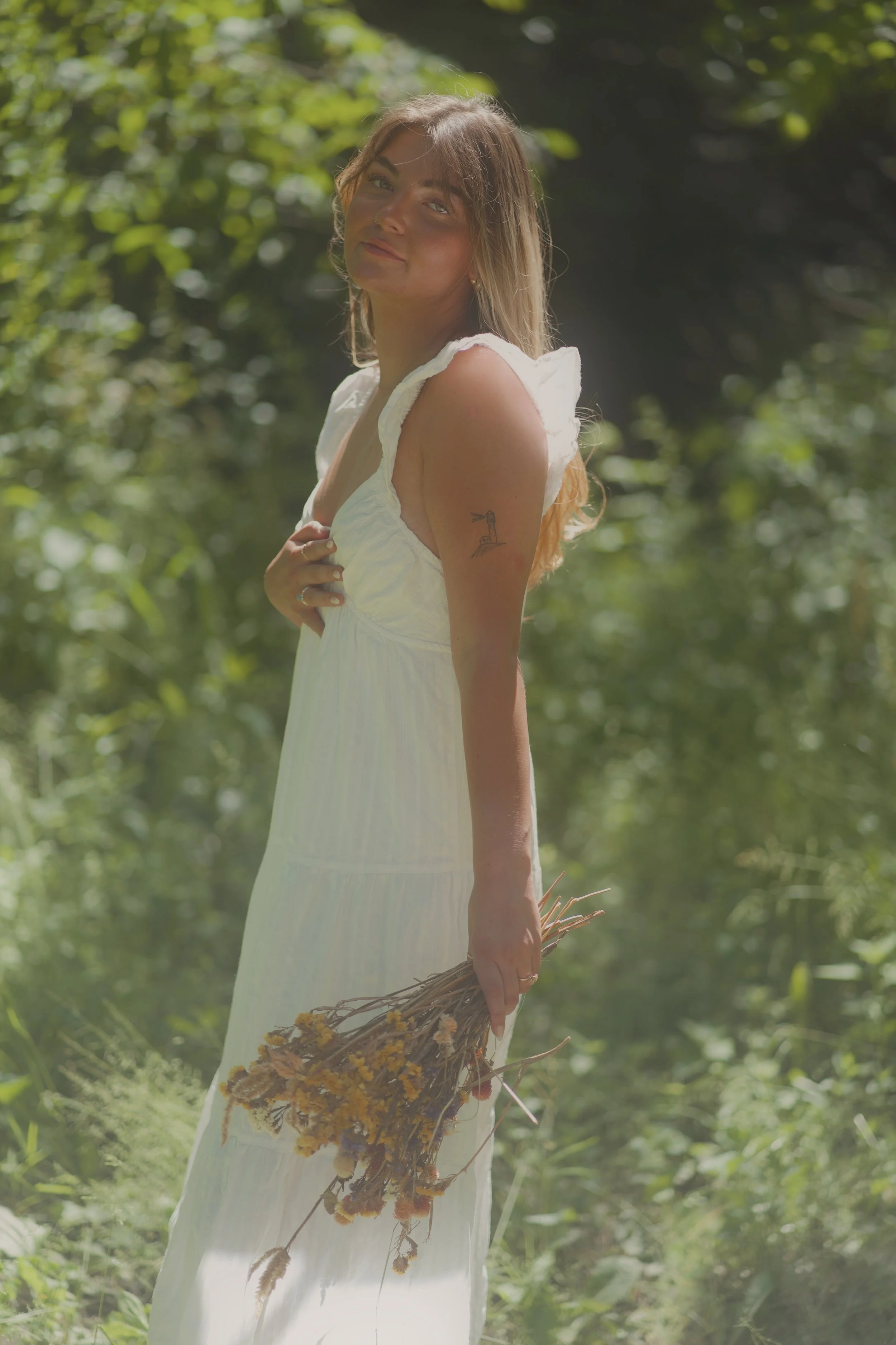 A young woman with light brown hair and blue eyes standing outdoors in a green, sunlit forest. She is wearing a white sleeveless dress and holding a small bundle of dried yellow flowers and stems in her right hand. She is looking at the camera with a slight smile and has a small tattoo of a lighthouse on her upper left arm.