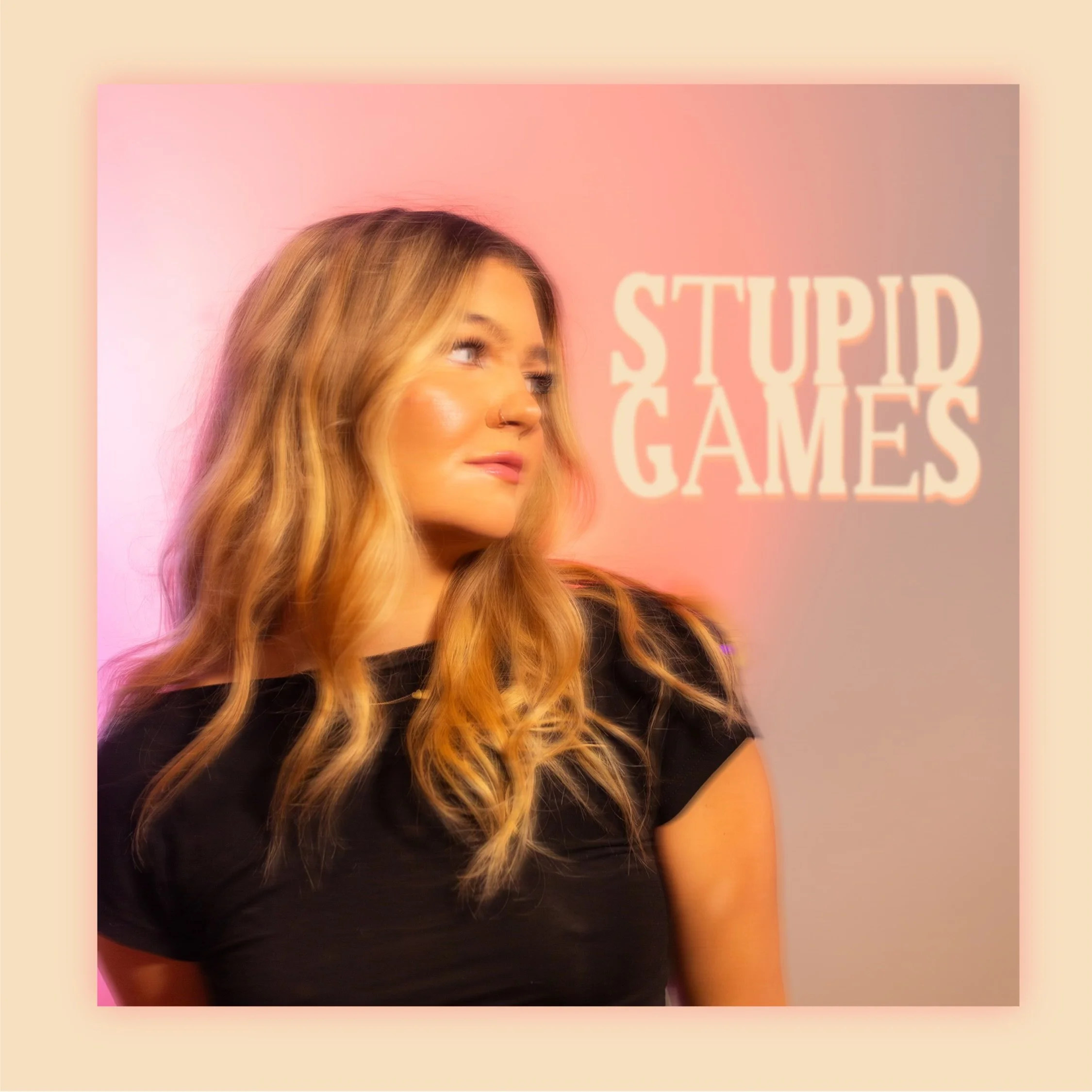 A woman with long, wavy blonde hair and light skin, wearing a black shirt, looking to the side against a pink background with partially visible white text.