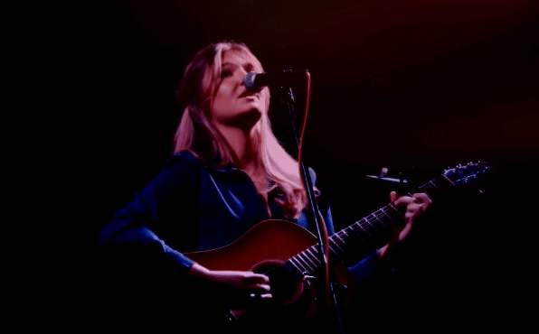 Young woman playing an acoustic guitar and singing into a microphone on stage, with a dark background and stage lighting.