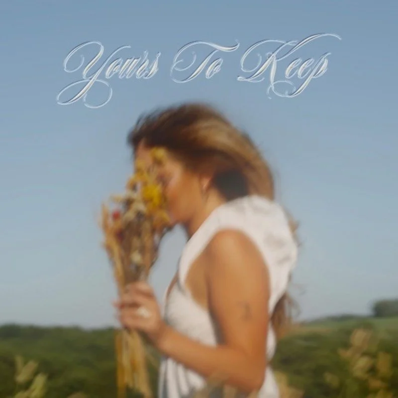 Blurred image of a woman with long hair holding a bouquet of flowers outdoors, with a landscape background and the text 'Yours To Keep' in cursive at the top.