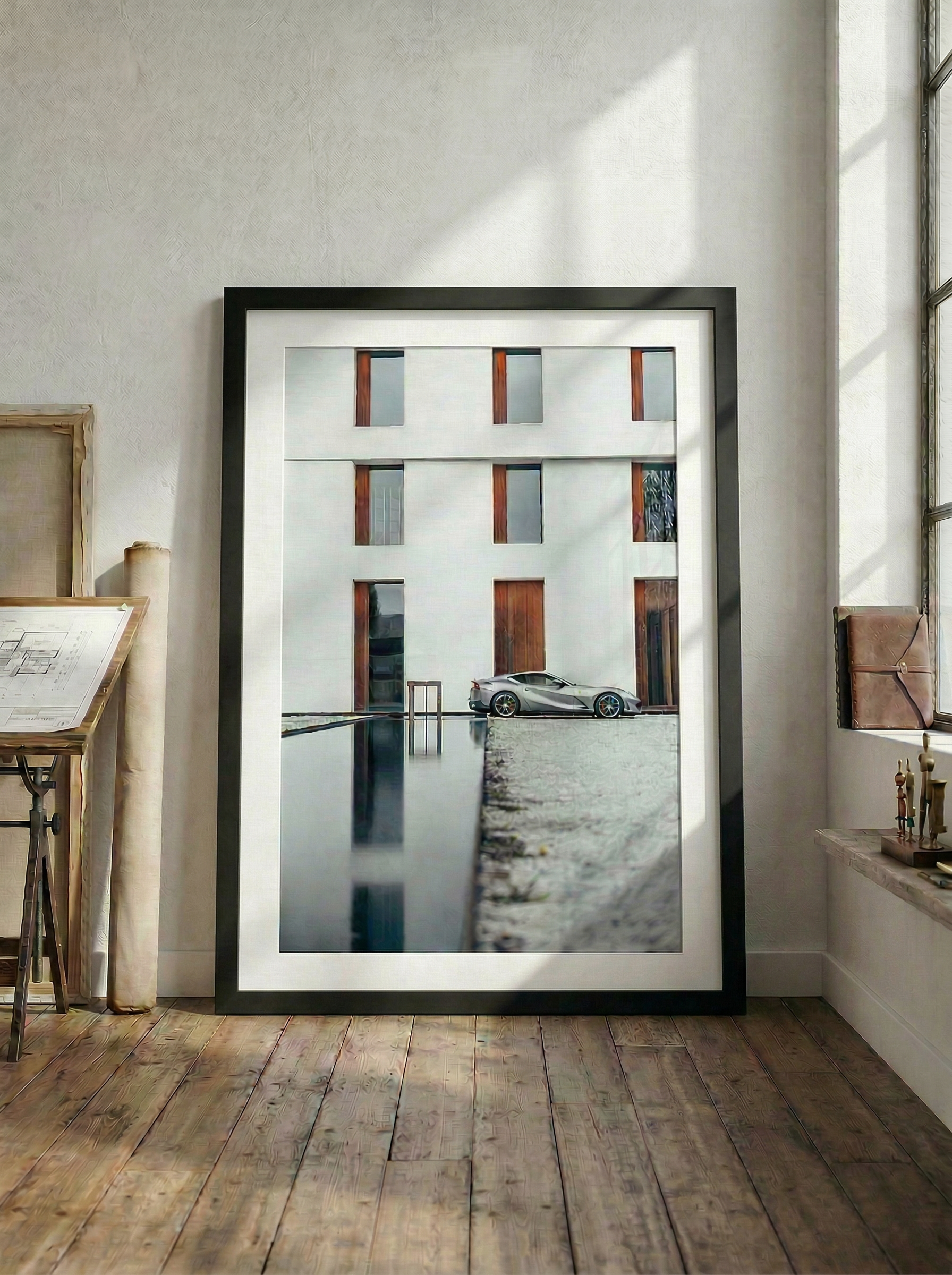 A framed photograph of a modern white building with tall vertical windows and a black sports car parked in front, leaning against a curb, leaning against a shallow water feature, with a wooden-desk and papers on the left and a window with a ledge on the right.