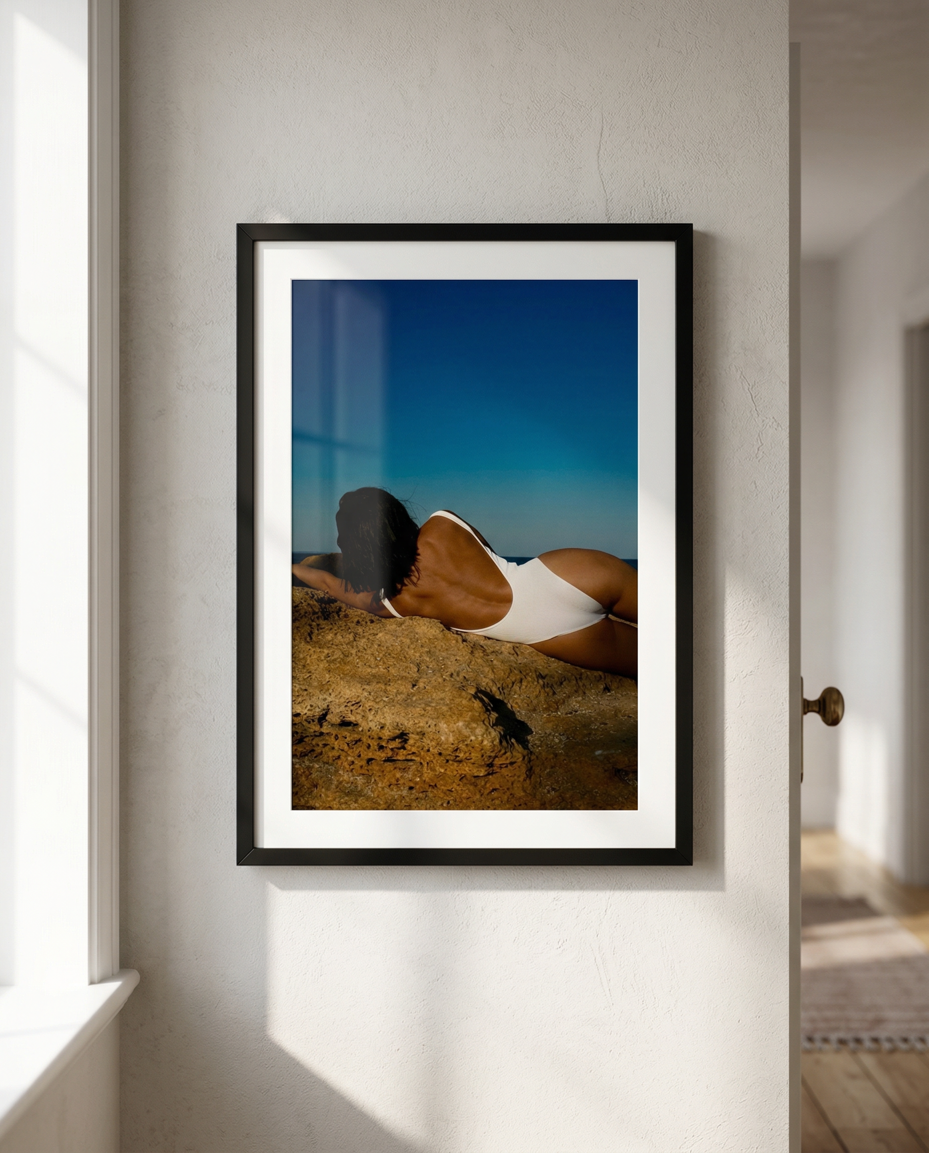 A framed photograph of a woman with dark hair lying on a large rock at the beach, wearing a white swimsuit, with the ocean and blue sky in the background, hanging on a white wall.