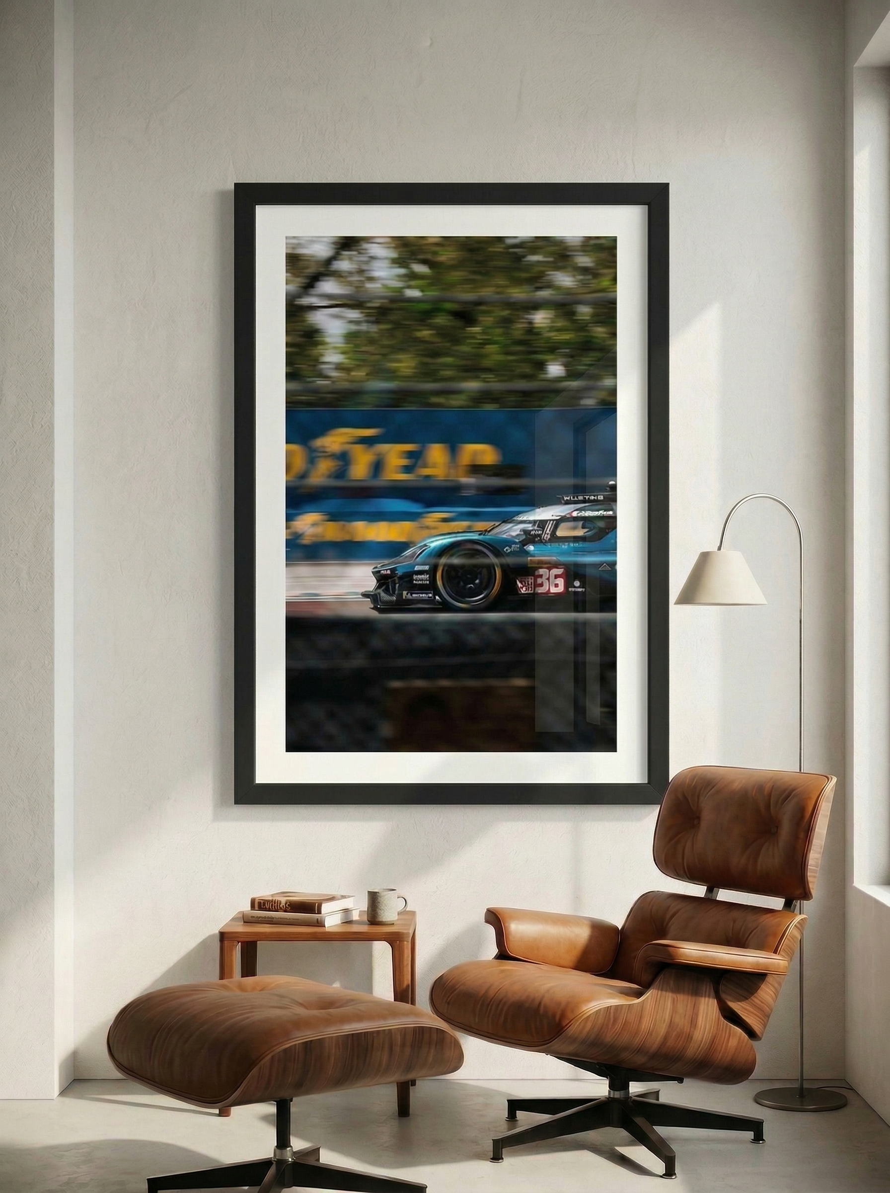 A framed photograph of a race car in motion hangs on a white wall in a modern living room with a brown leather chair and matching ottoman, a small wooden side table with books and a mug, and a floor lamp.