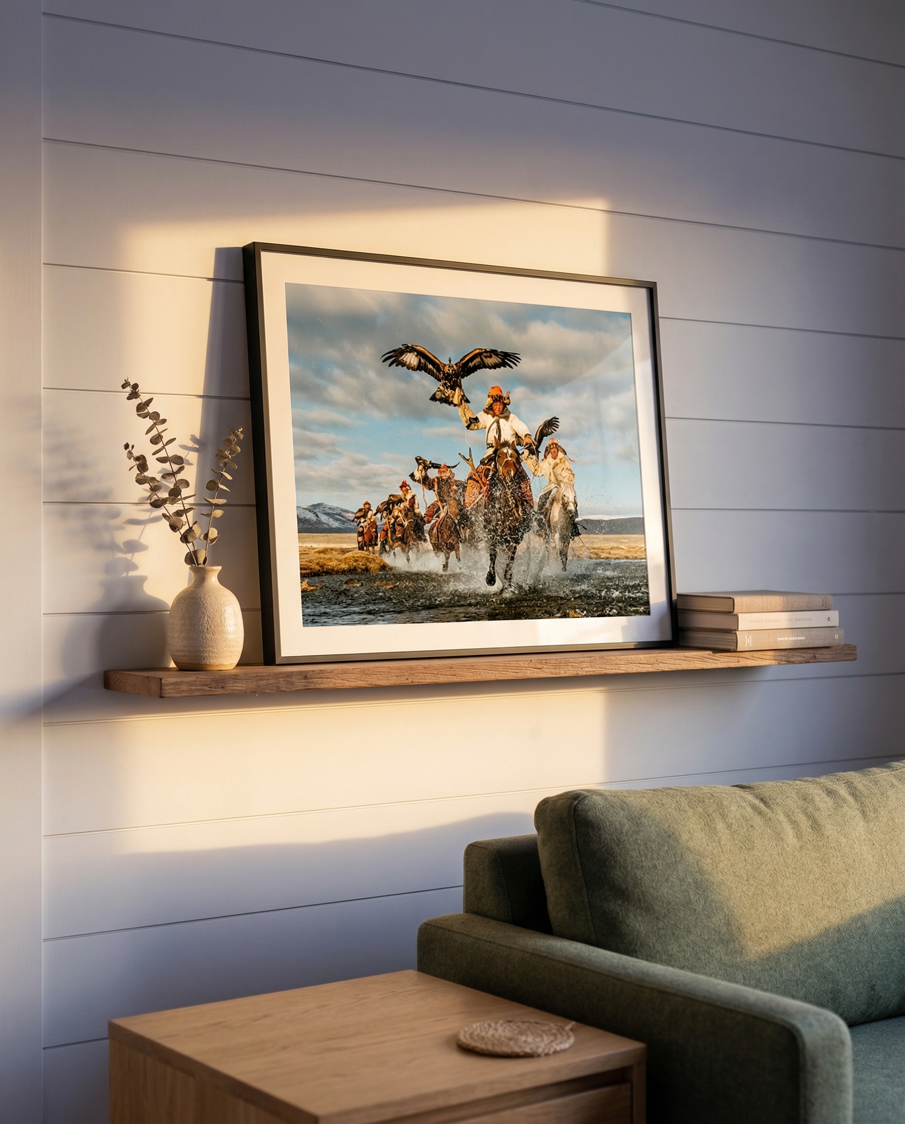 A framed picture of people on horseback herding cattle in a desert landscape, with mountains and a cloudy sky in the background, hung on a white wall with horizontal panels. There is a small vase with eucalyptus branches on a wooden shelf to the left of the picture and a stack of books to the right. A portion of a green sofa and a wooden side table with a coaster are also visible.