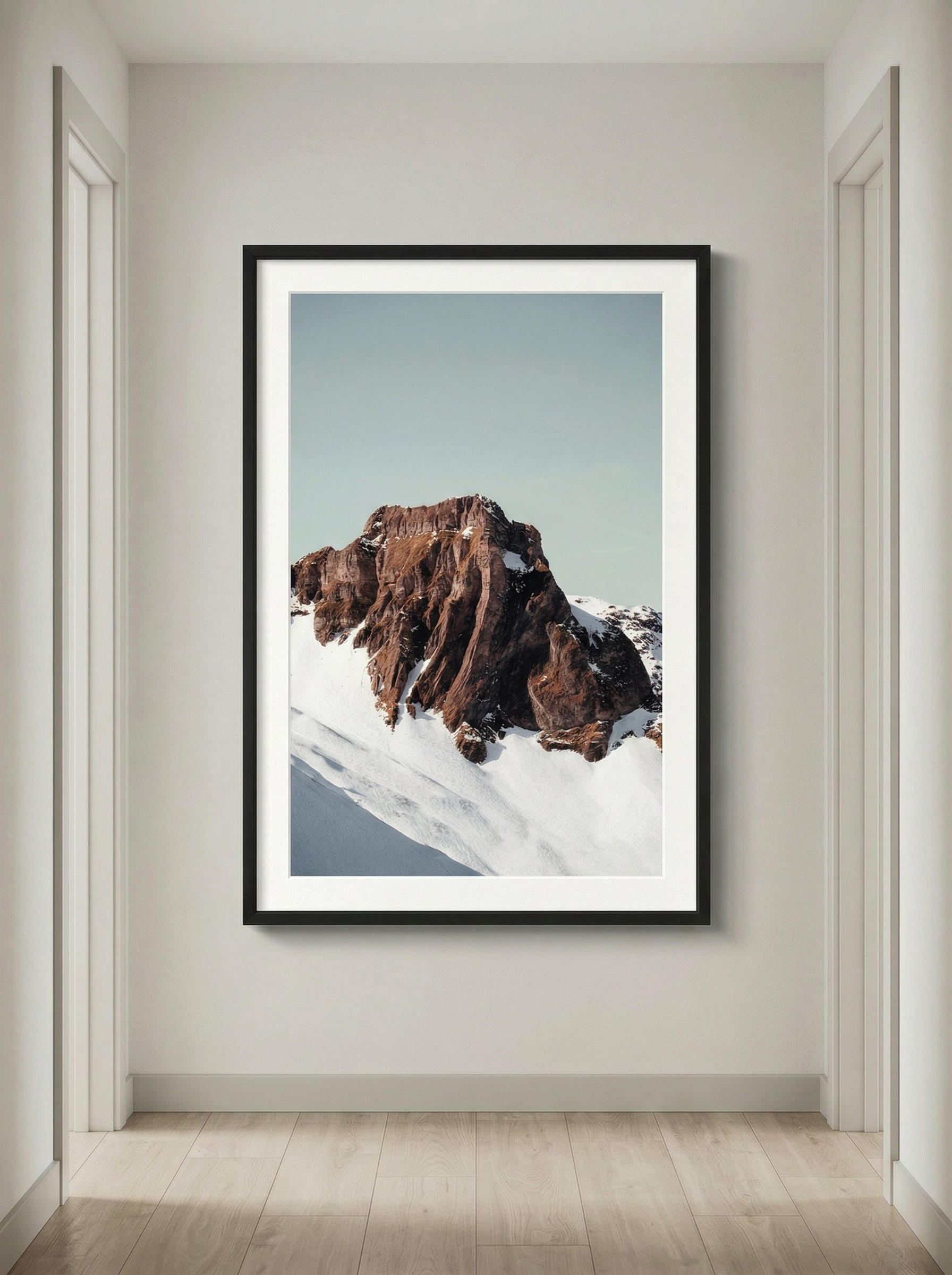 Framed photograph of a mountain with snow and rocky peaks, hung on a white wall in a hallway with light wood flooring.