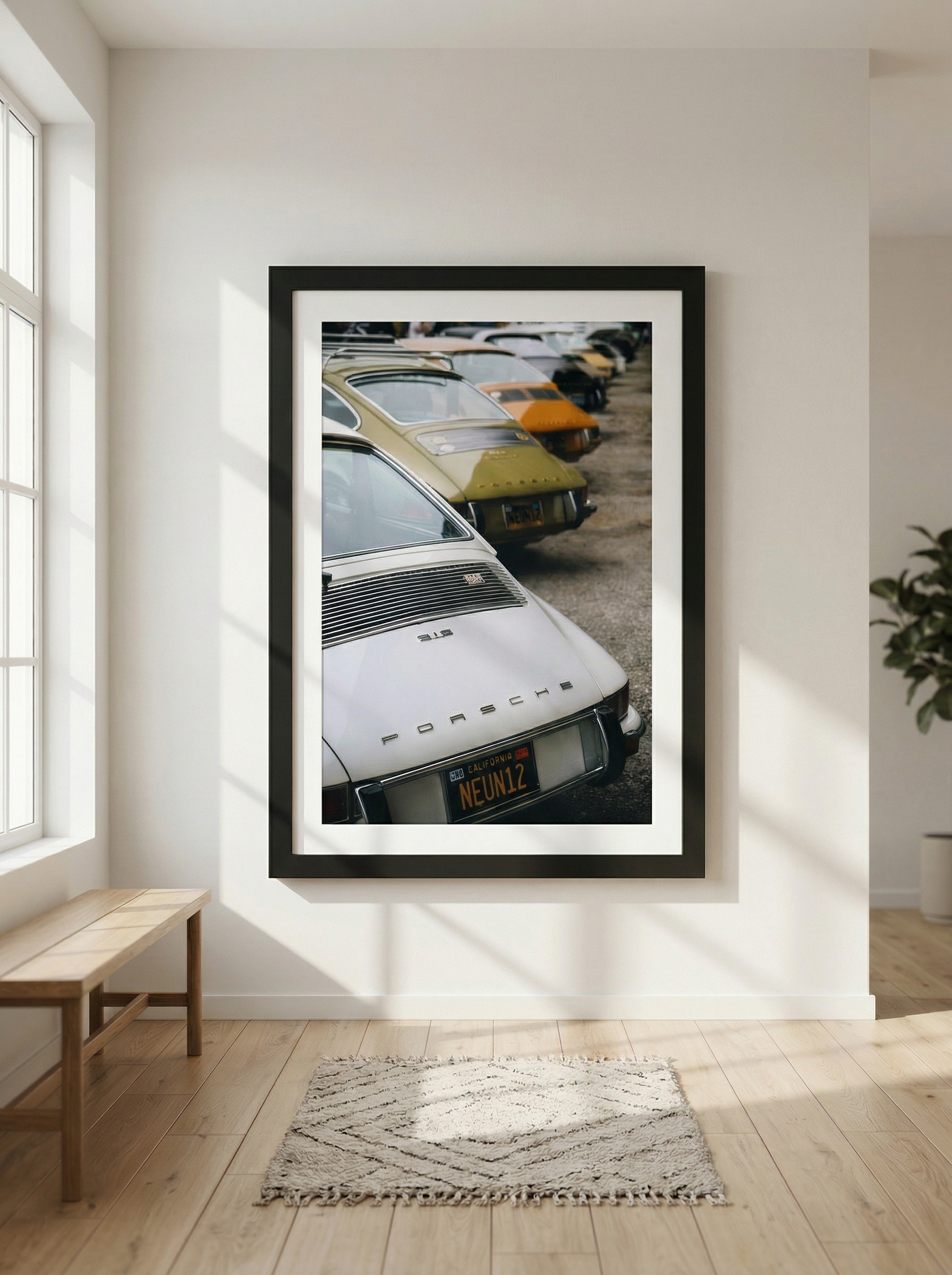 A framed poster of a lineup of classic Porsche cars with a focus on the front of a silver Porsche with a California license plate reading 'NEUN12' hangs on a white wall in a bright room with wooden flooring, a small wooden bench, a patterned rug, and a potted plant.