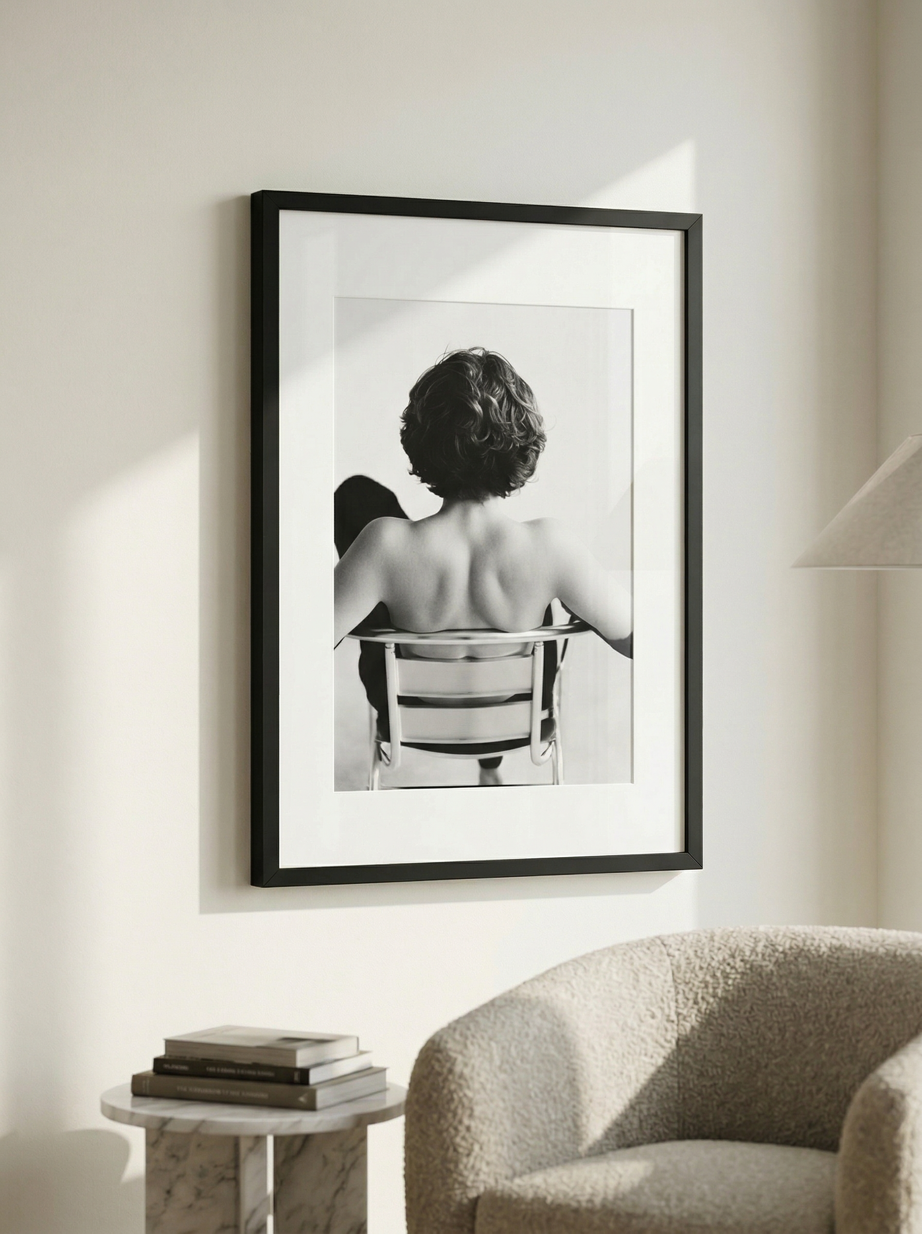A black-and-white photograph of a woman with short curly hair viewed from behind, seated on a chair, hanging on a white wall in a modern interior.