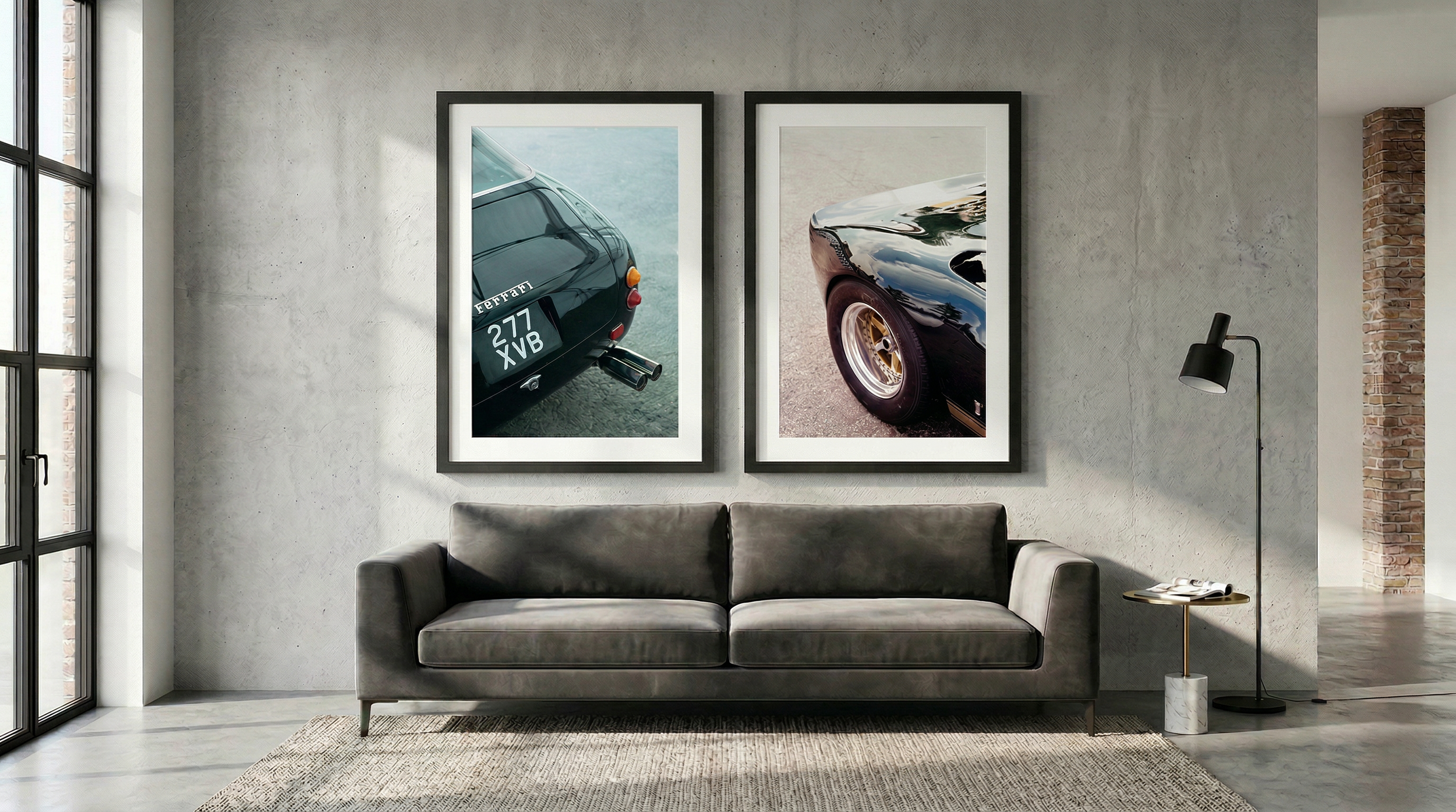 Modern living room with concrete walls, large windows, and a dark gray sofa. Two framed photographs of vintage sports cars hang on the wall behind the sofa. A black floor lamp stands beside a small side table, which holds a book and a cup.