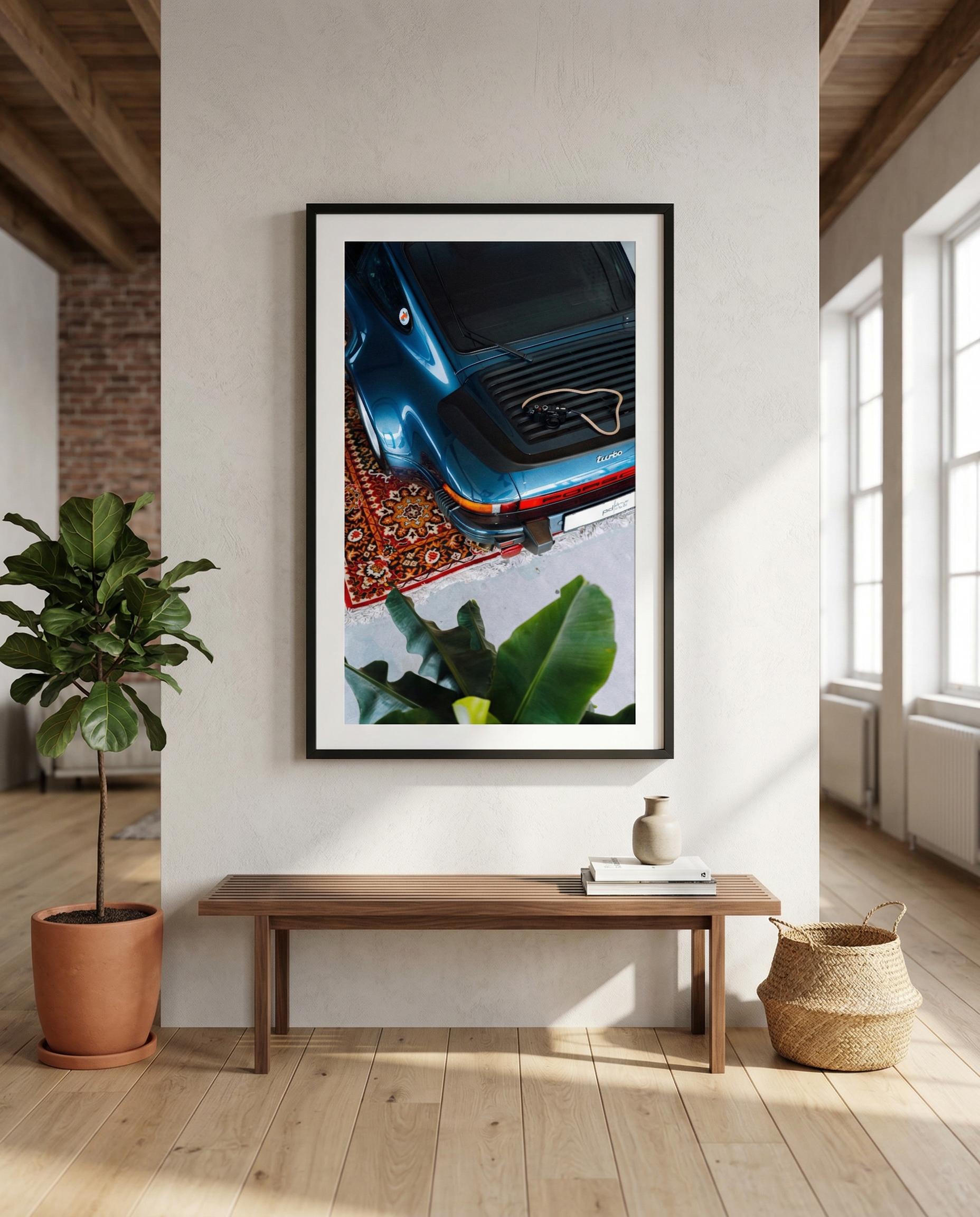 Framed photo of a blue vintage Porsche 911 Turbo with a plant in the foreground, in a minimalist interior with wooden flooring, a wooden bench, a potted plant, a book with a small vase, and natural light from large windows.