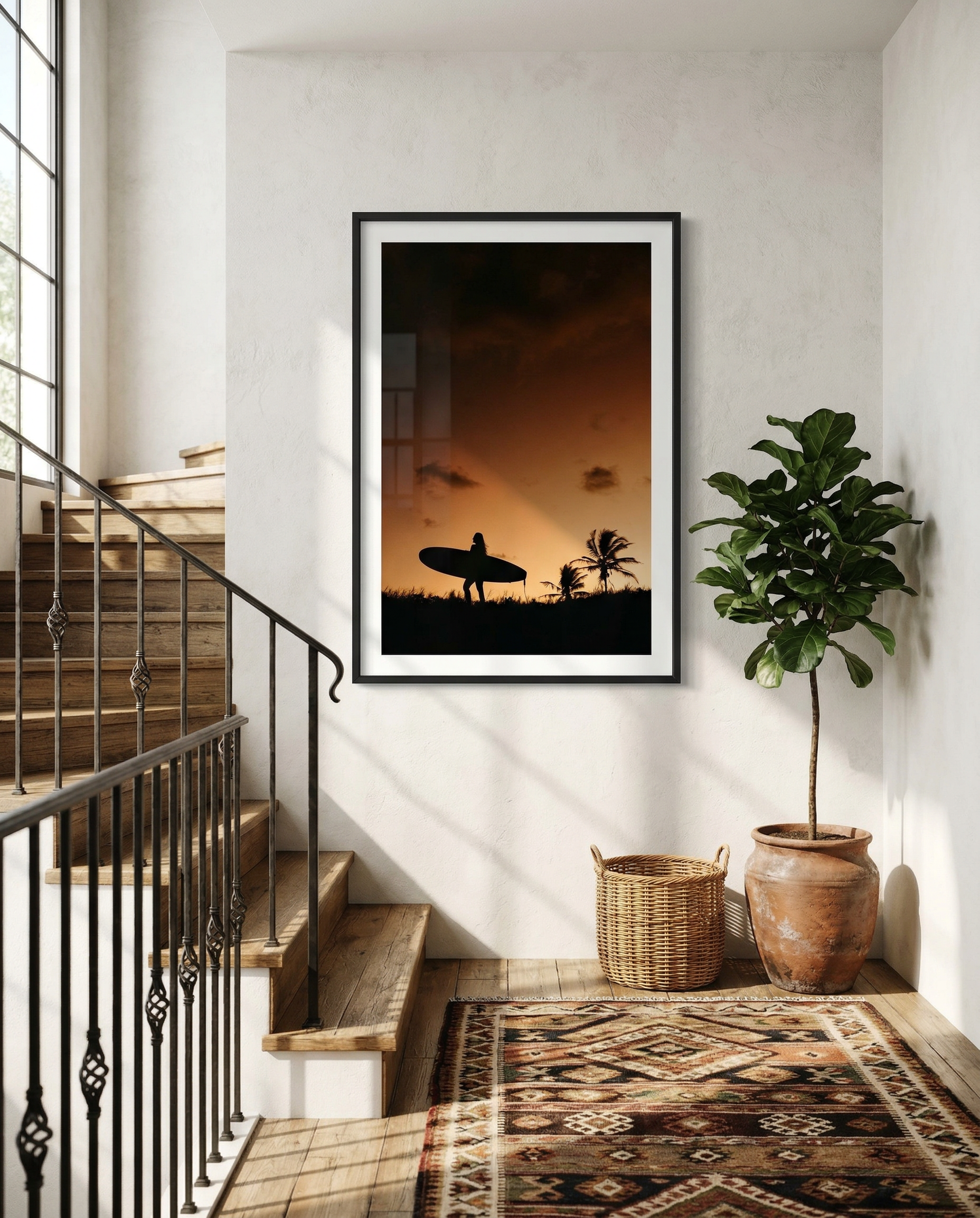 Interior view of a hallway with a framed sunset photo of a person with a surfboard near palm trees, a potted plant, a woven basket, a patterned rug, and wooden stairs with a iron railing.