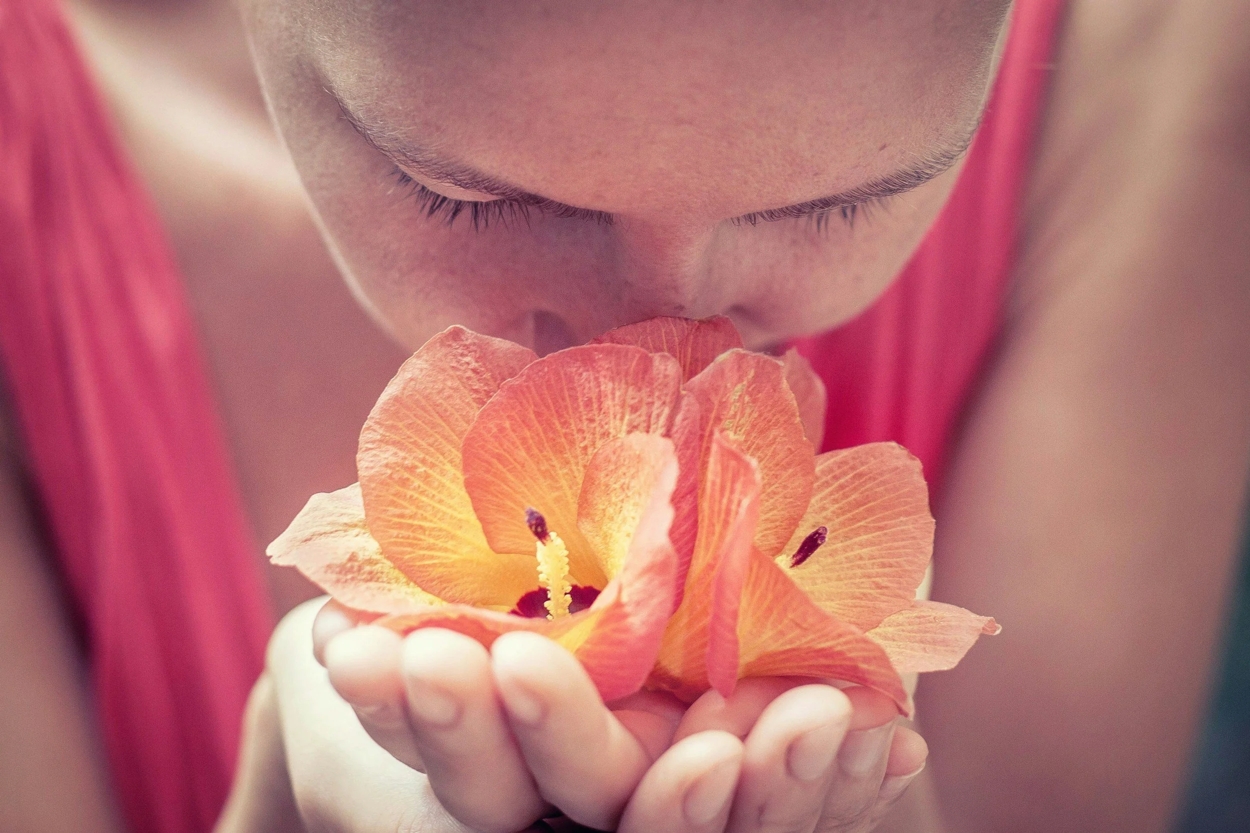 A person holding a bright orange flower close to their face while smelling it, with their eyes closed.