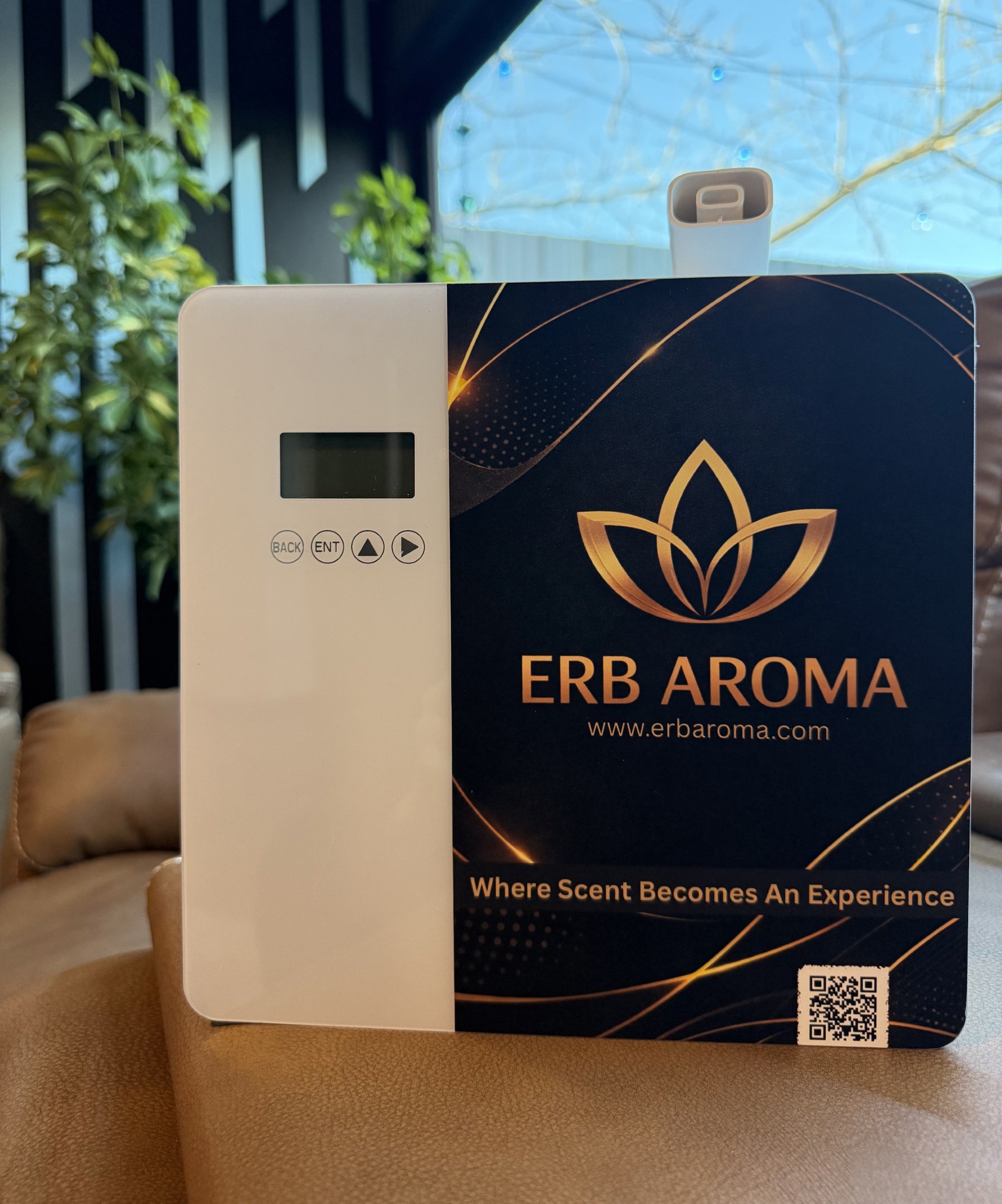 A wellness or aroma diffuser device branded ERB AROMA with a digital display and control buttons, placed on a tan surface, with a dark blue information card with gold accents and logo.