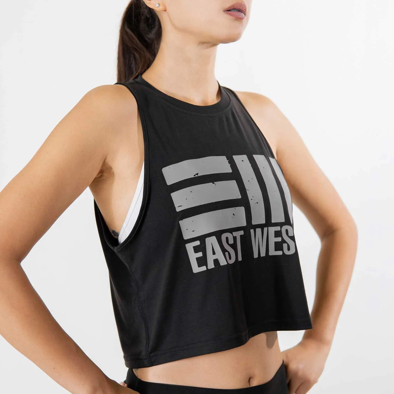 A woman wearing a black sleeveless sports top with the words 'EAST WEST' printed on the front, standing with her hand on her hip against a plain white background.