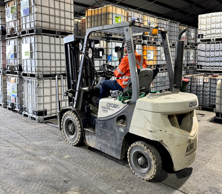 A worker in an orange safety vest driving a Crown forklift in a warehouse filled with large plastic containers stacked on metal pallets.