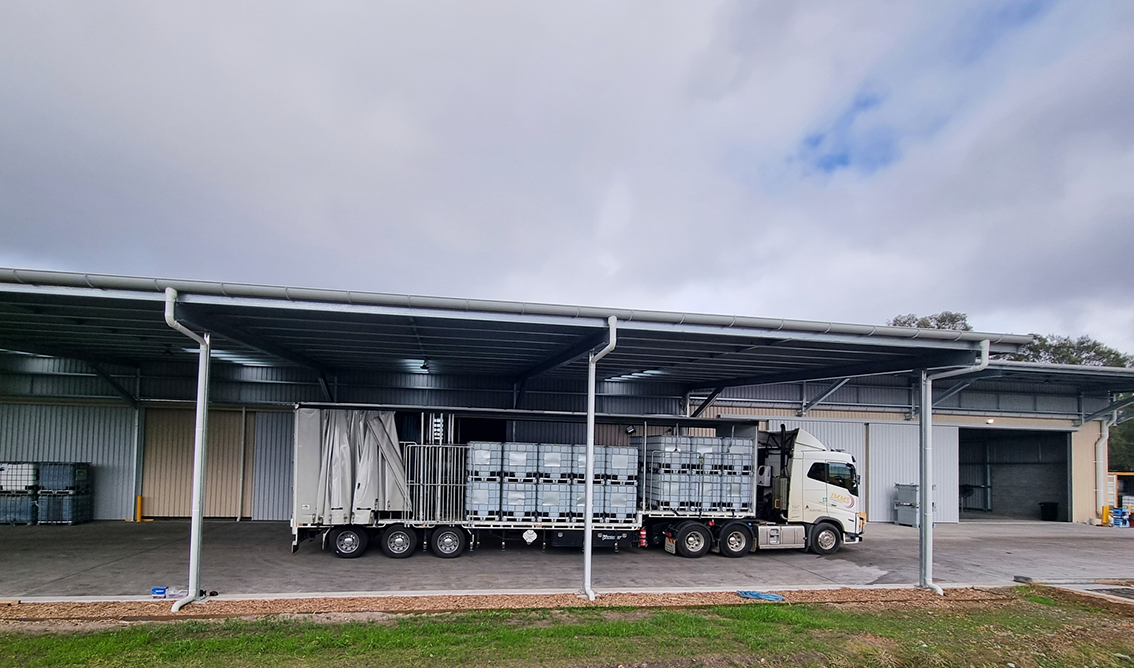 A semi-truck parked under a large metal canopy at a warehouse loading dock, with pallets of large bottles or containers loaded onto the truck.