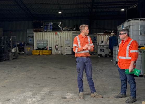 Two men in orange safety vests and jeans standing and talking inside a warehouse with industrial equipment and storage containers.