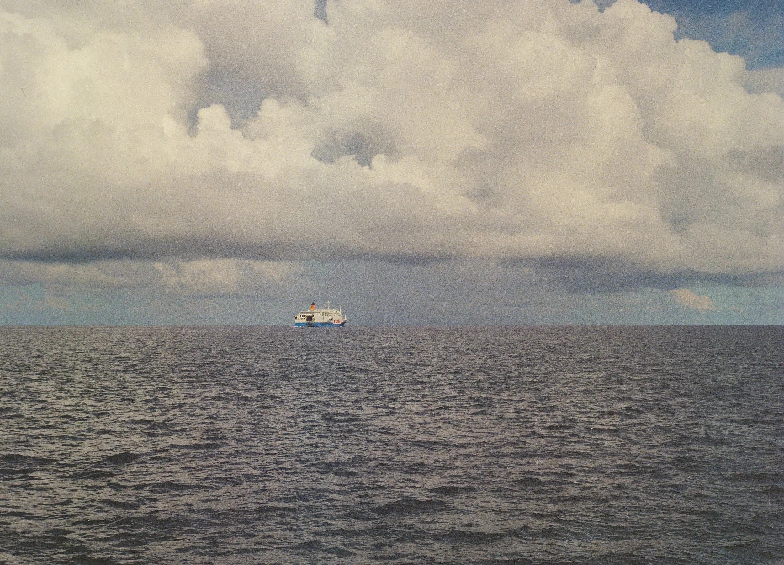 A ship is floating on a vast, calm ocean under a cloudy sky.