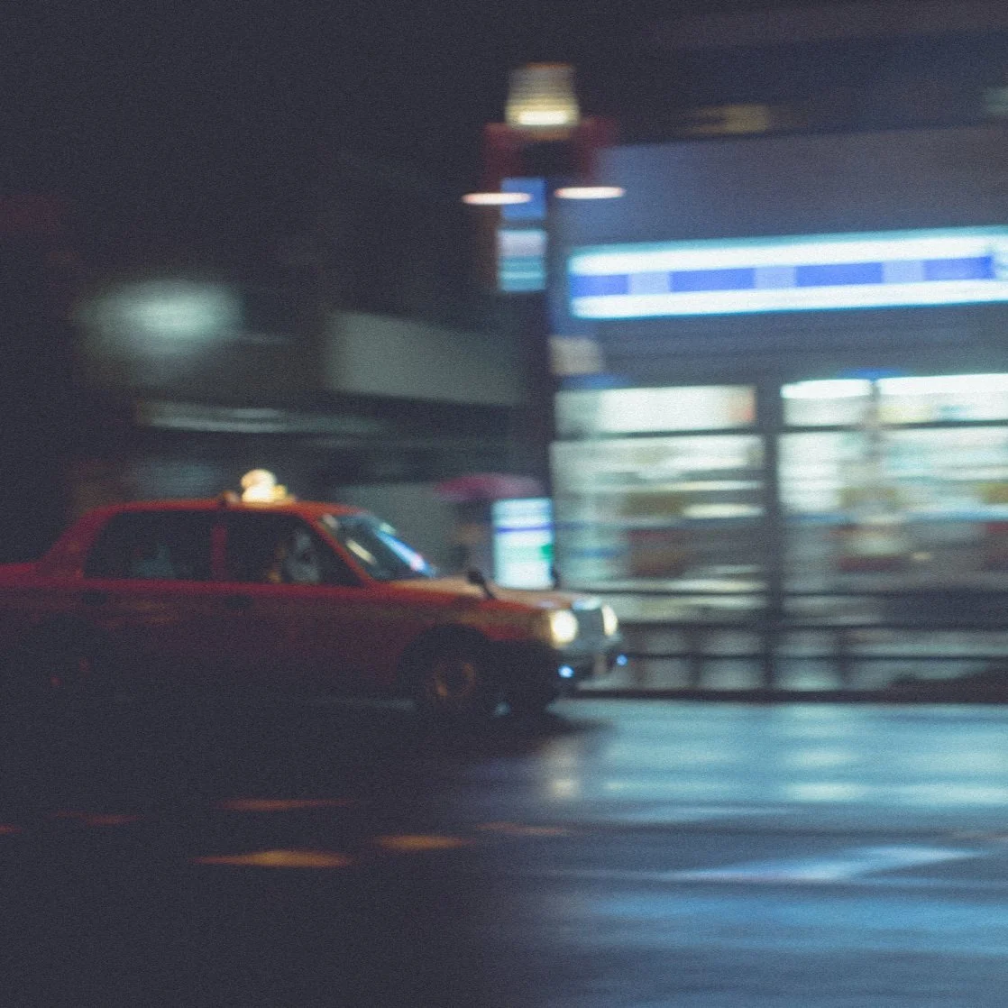 A blurred image of a taxi cab driving at night with city lights and a large sign in the background.