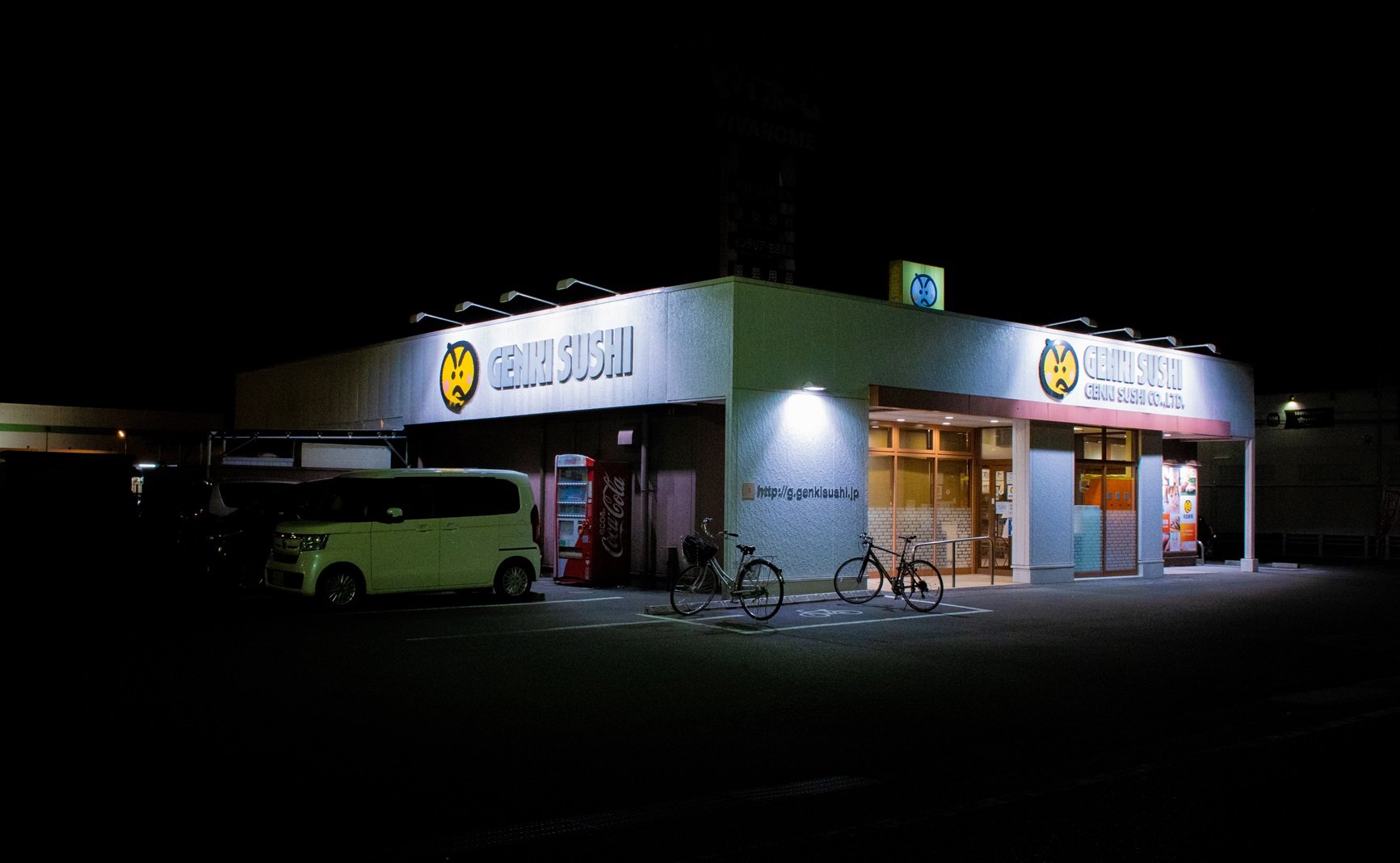 Night view of Geneki Sushi restaurant with illuminated signage, bicycles parked outside, and a vending machine, in a dimly lit parking lot.
