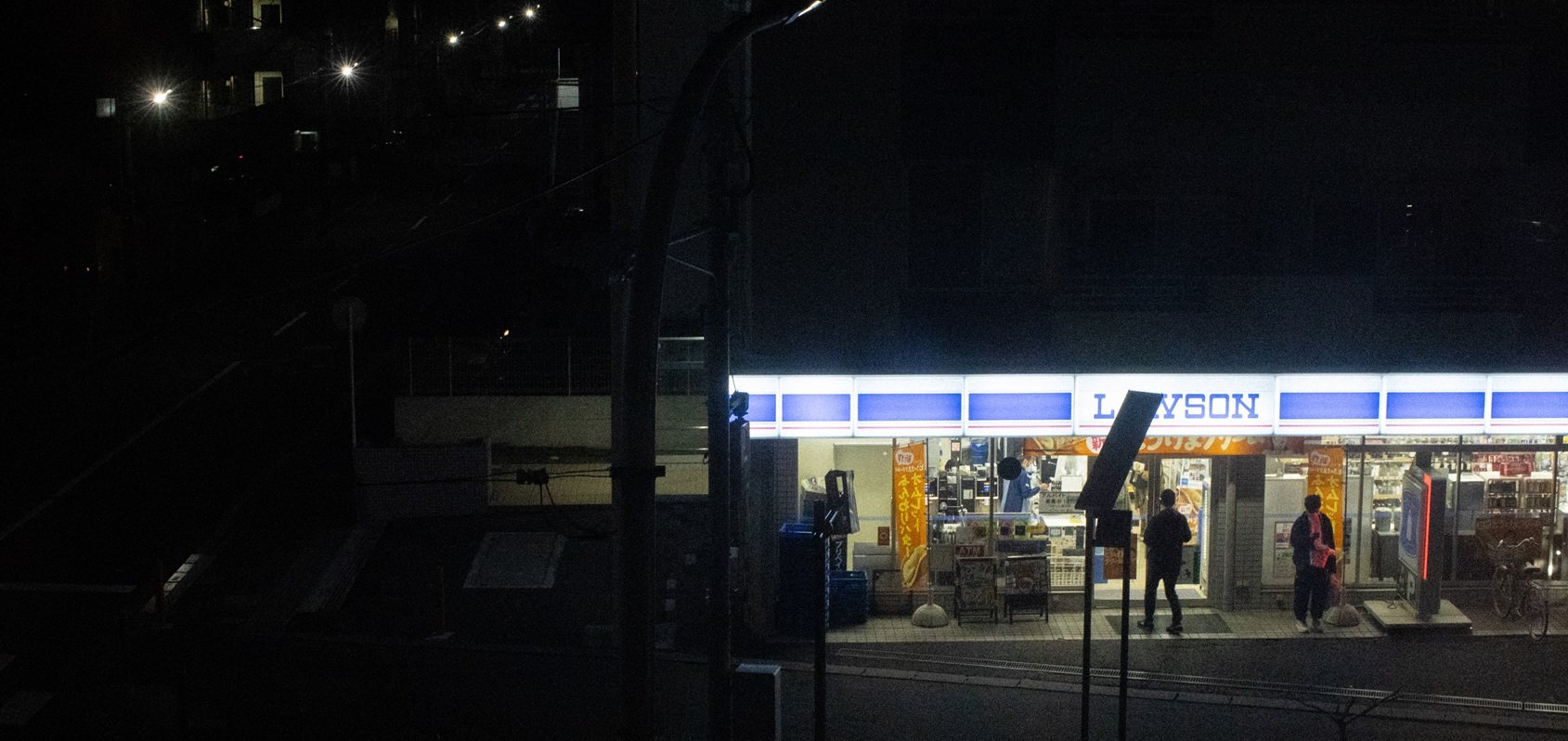 Nighttime scene of a Lawson convenience store with a lit sign outside. Three people are near the entrance, and others are inside the store. A bicycle is leaning outside the store, and some shopping carts are visible.