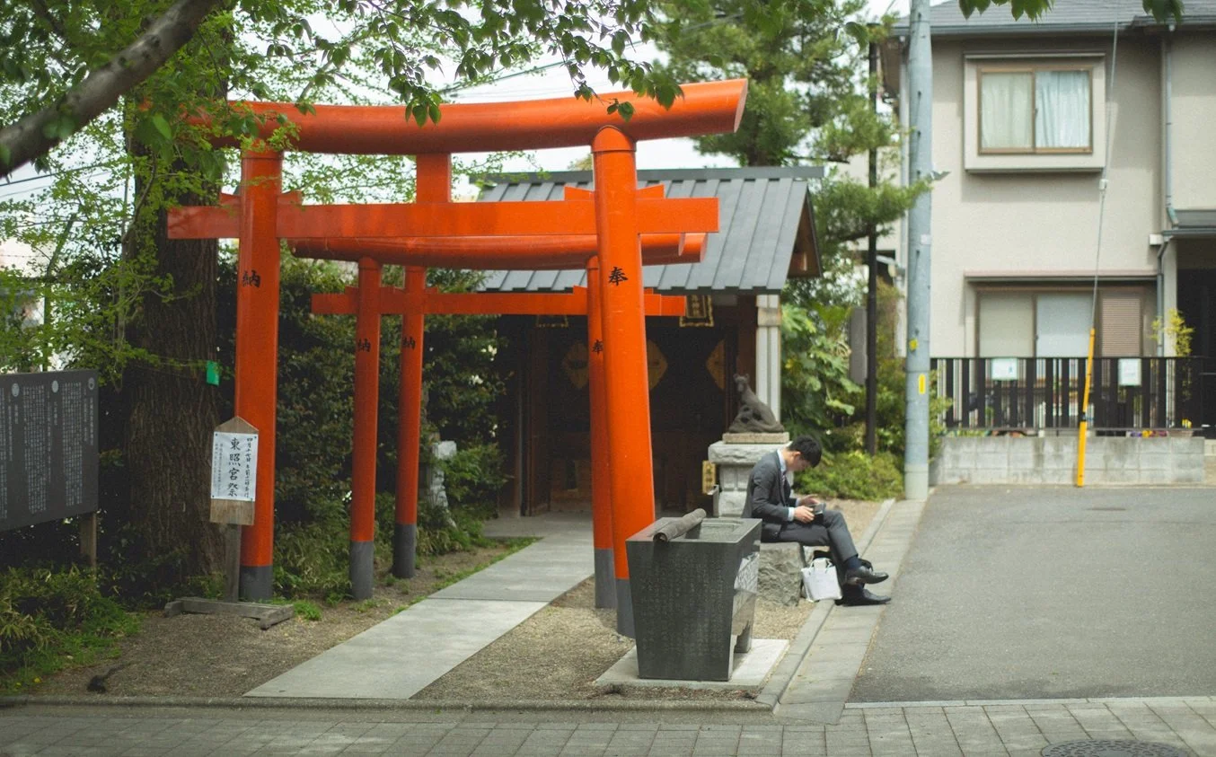 A man in business attire sitting on a stone bench outside a small Shinto shrine with a red torii gate, surrounded by trees and nearby buildings.